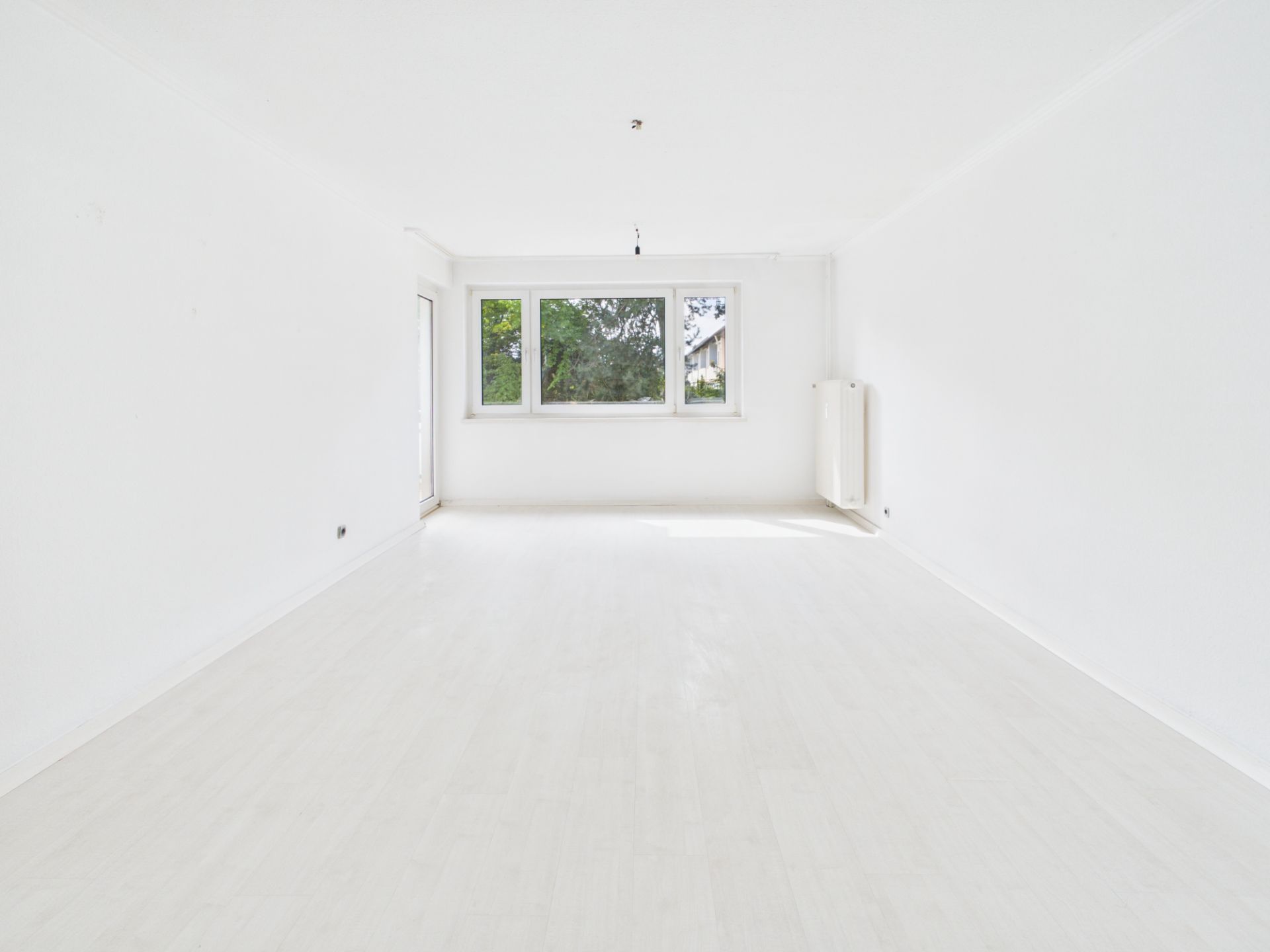 Helles, leeres Zimmer mit weißen Wänden, großem Fenster und Heizkörper an der rechten Wand.