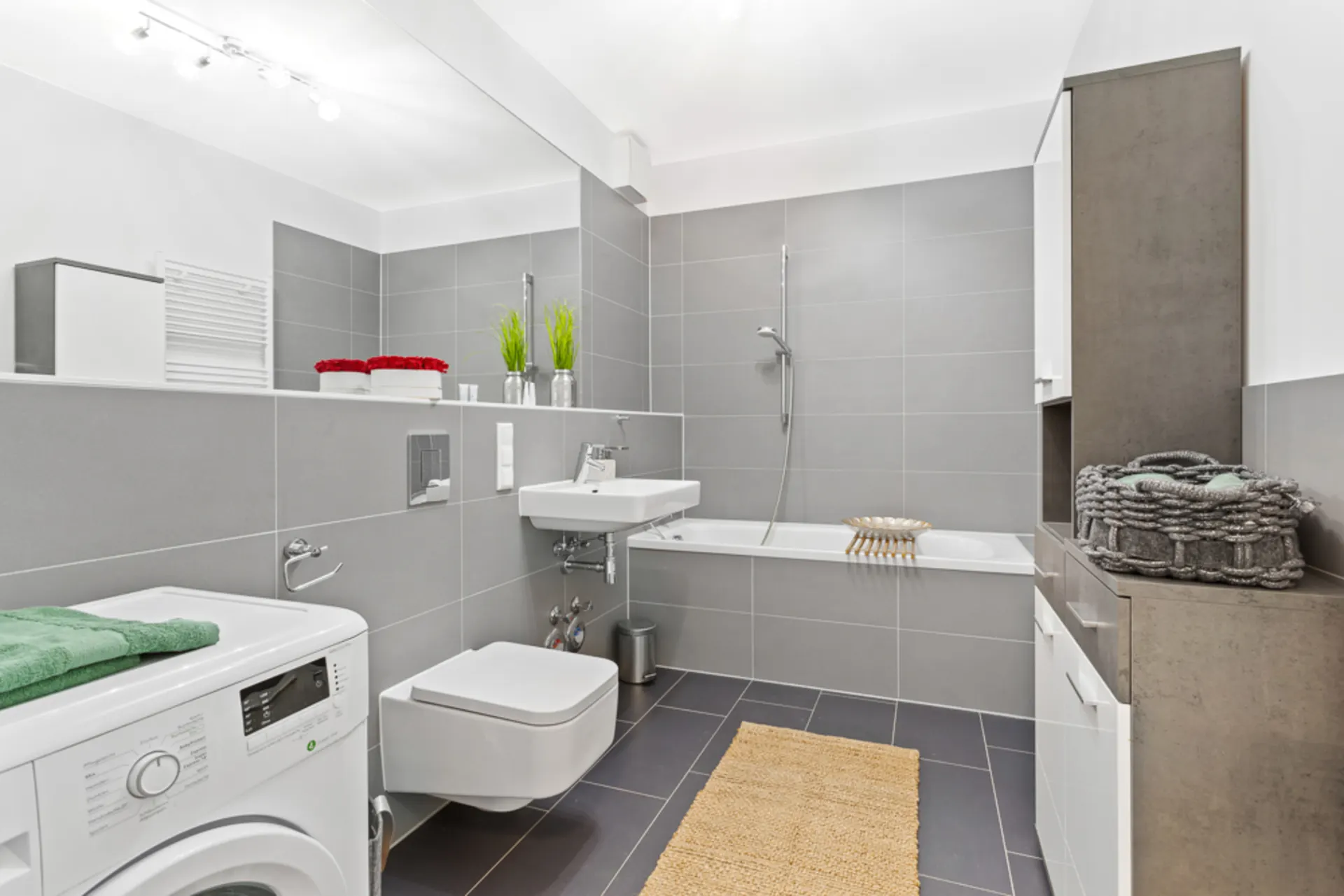 Modernes Bad mit grauen Fliesen, Waschmaschine, Badewanne, Waschbecken und Regal.
