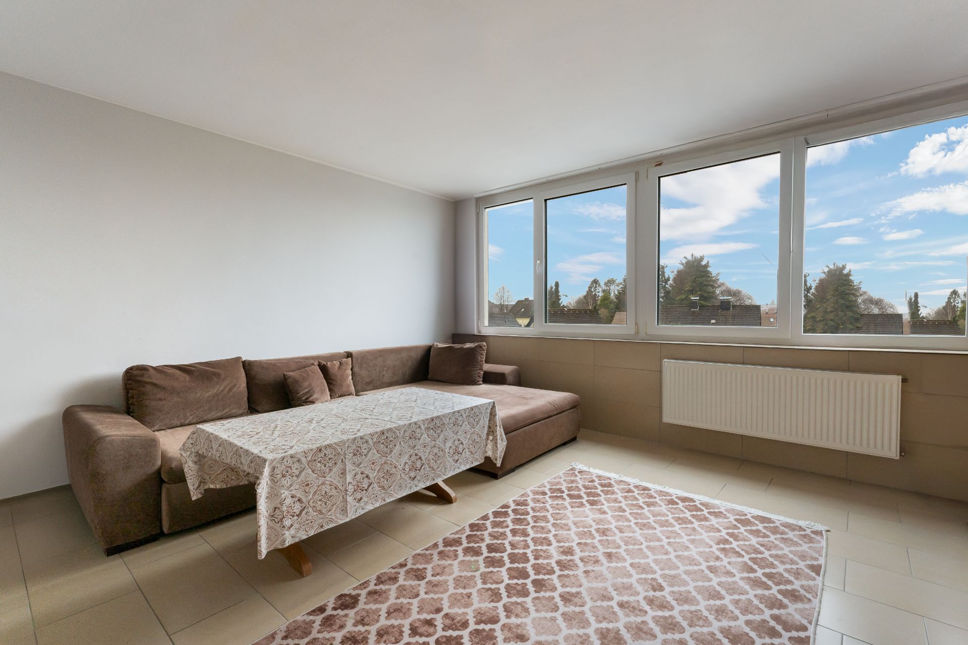 Wohnzimmer mit braunem Ecksofa, Tisch mit Tischdecke und großem Fenster.