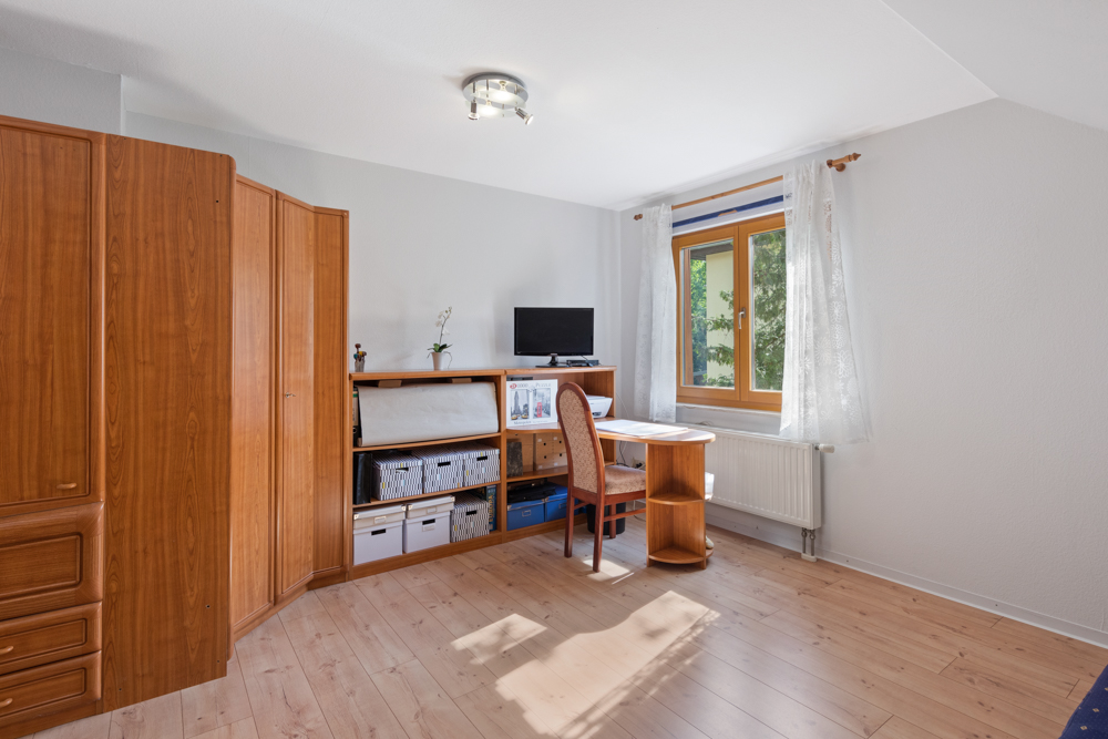 Helles Zimmer mit Holzschrank, Schreibtisch, Stuhl und Fenster mit weißen Gardinen.