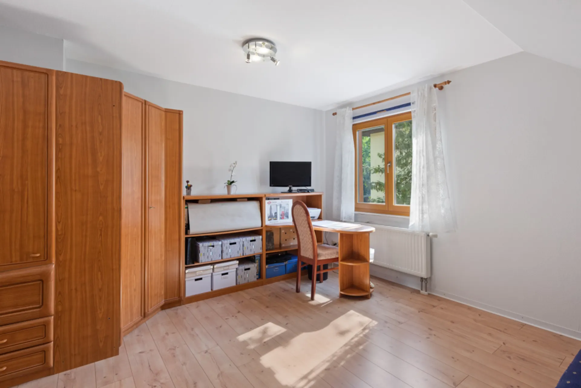 Helles Zimmer mit Holzschrank, Schreibtisch, Stuhl und Fenster mit weißen Gardinen.