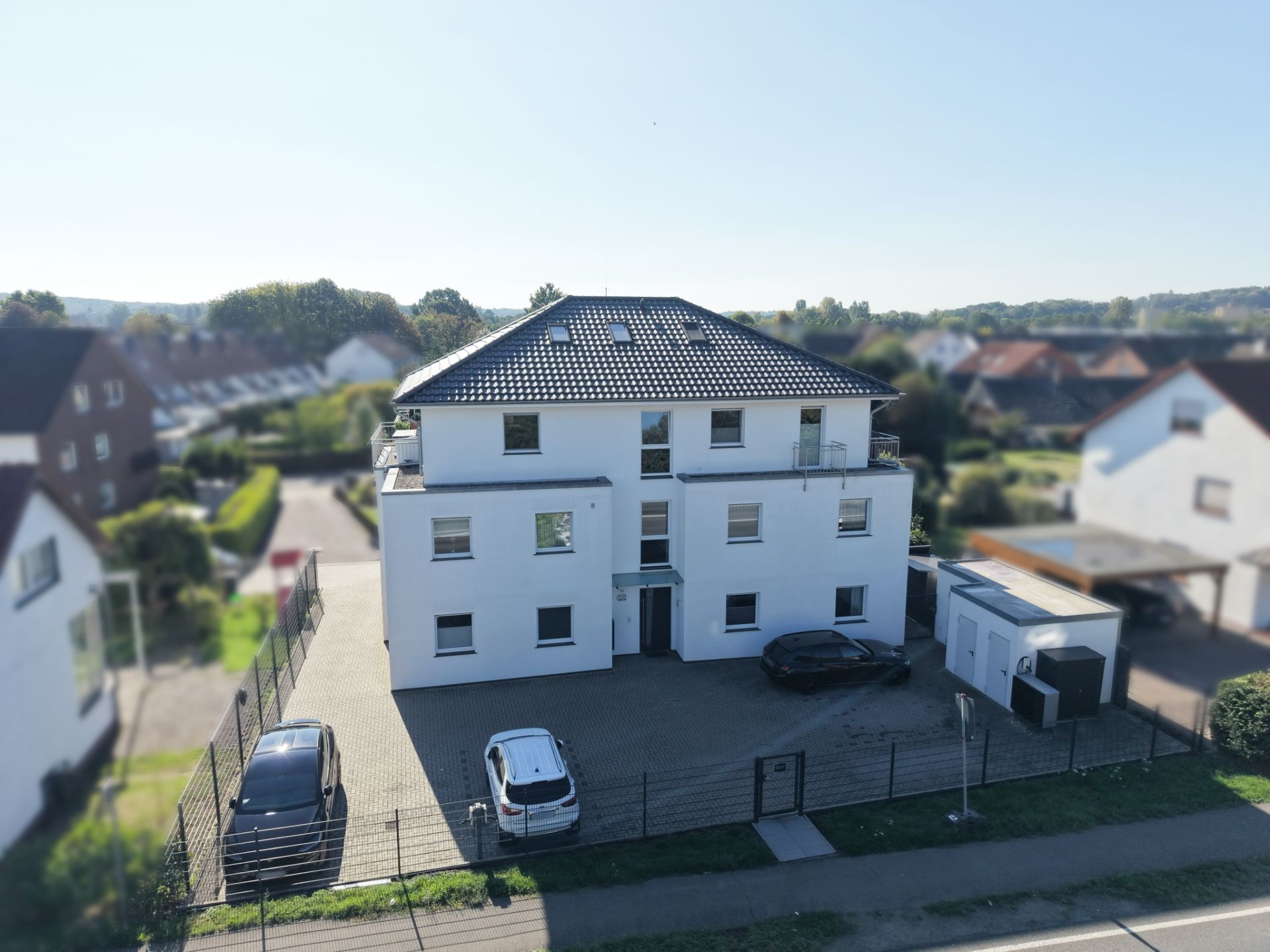 Modernes, weißes Wohnhaus mit drei Etagen, Parkplatz und zwei Autos davor.