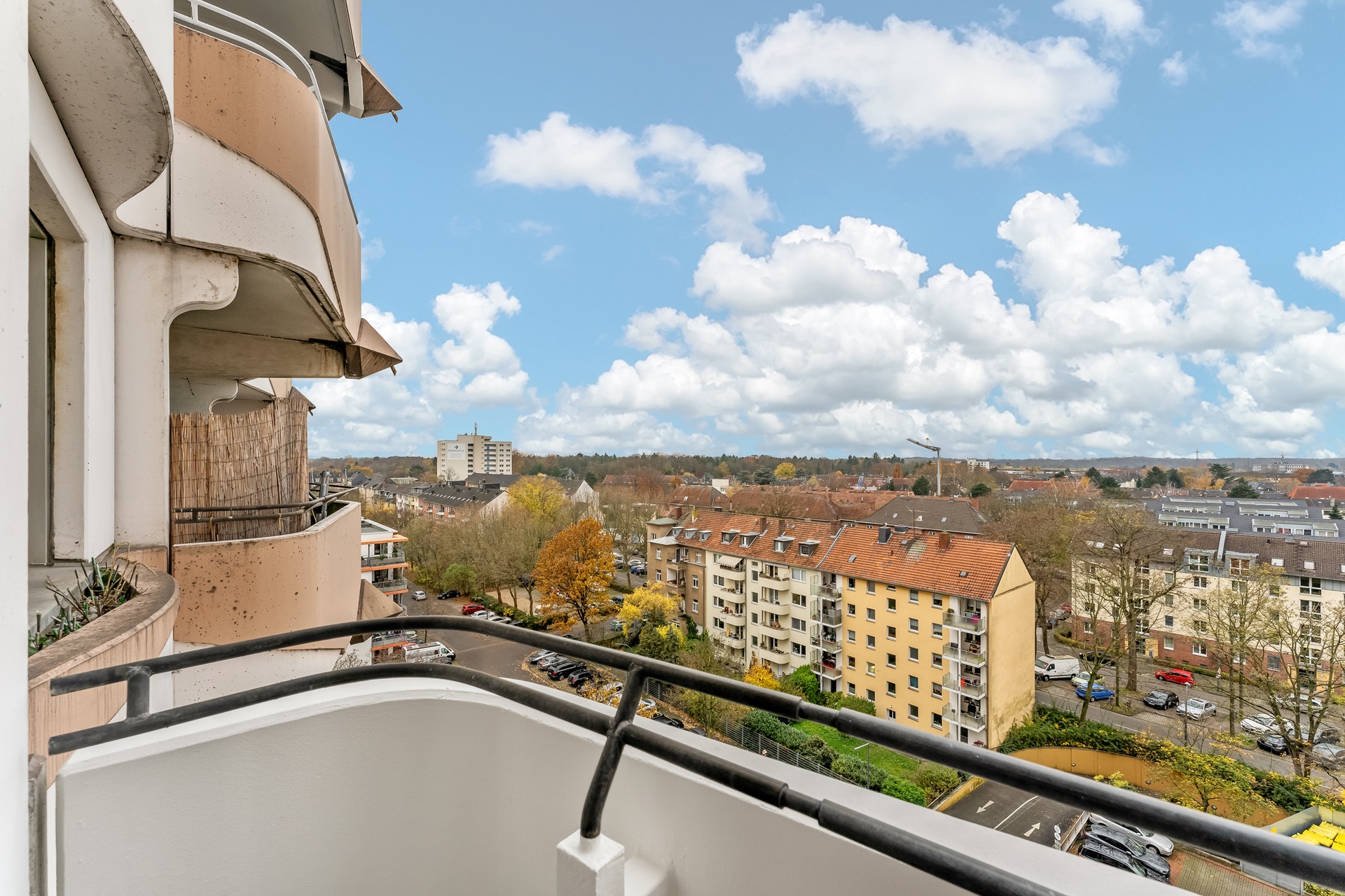 Blick von einem Balkon auf Wohnhäuser, Bäume und parkende Autos unter blauem Himmel.