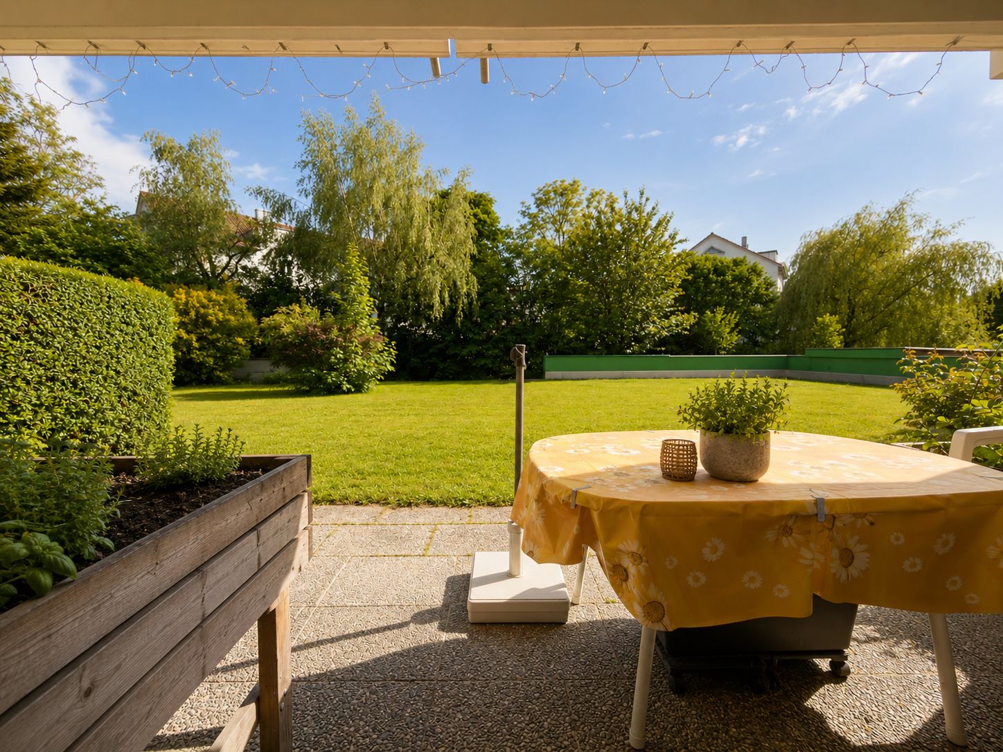 Terrasse mit gelbem Tisch, Hochbeet und Blick auf einen grünen Garten mit Bäumen.