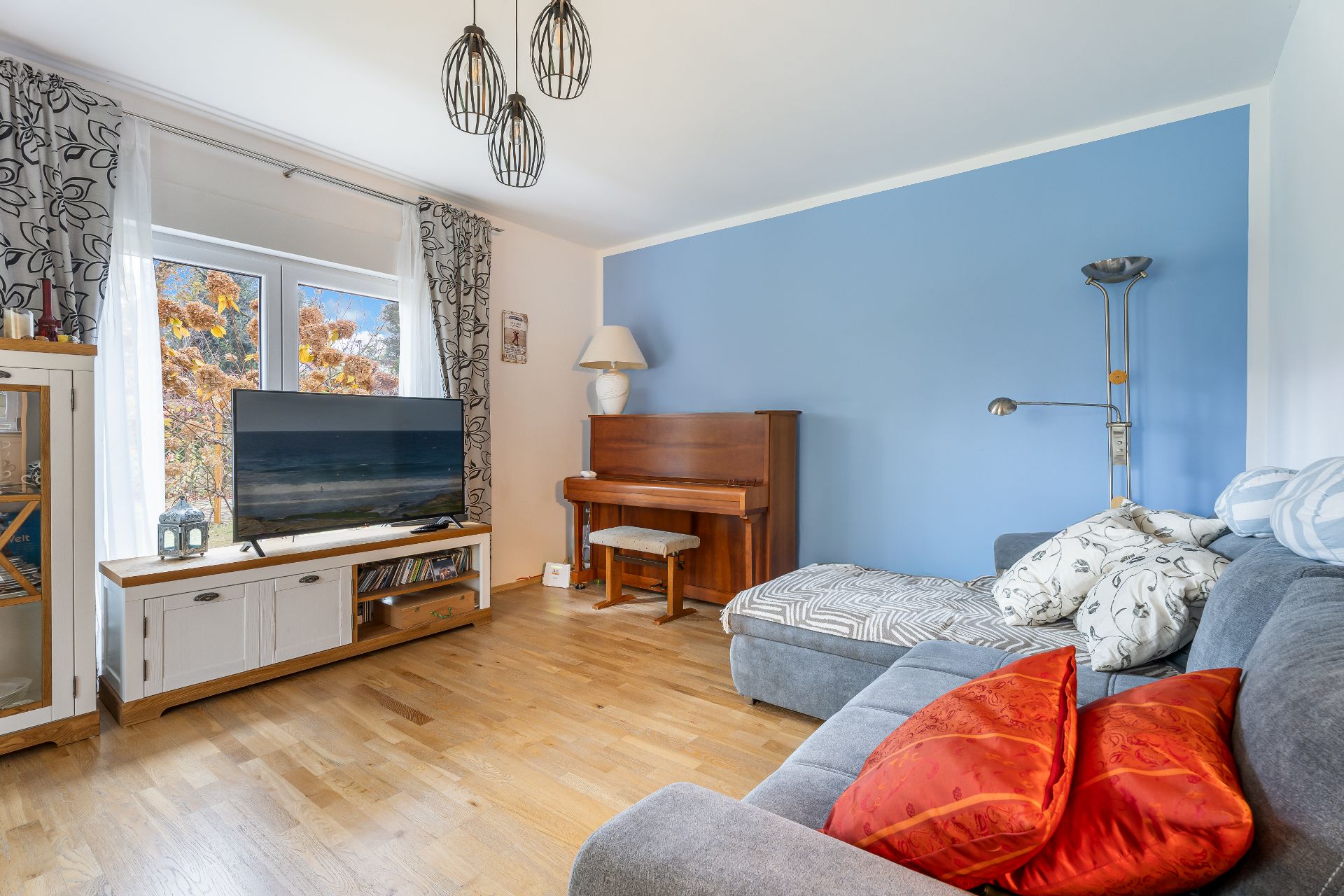 Wohnzimmer mit blau gestrichener Wand, Sofa, Klavier und Fernseher vor Fenstern.