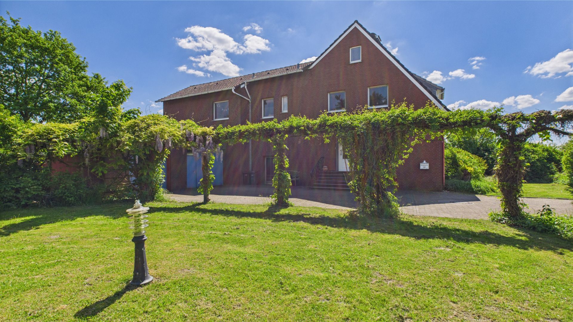 Backsteinwohnhaus mit bewachsener Pergola und großem Rasen im Vordergrund bei sonnigem Wetter.