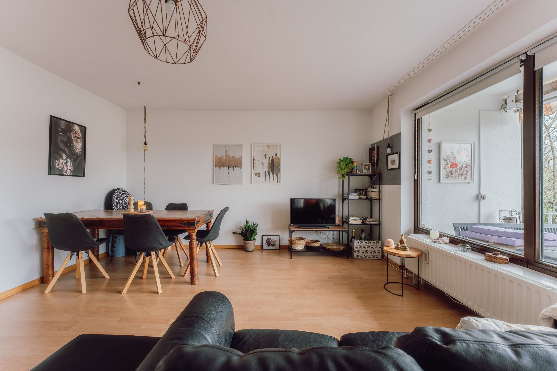 Helles Wohnzimmer mit Esstisch, schwarzen Stühlen, Sofa, Regal, Fernseher und großem Fenster.