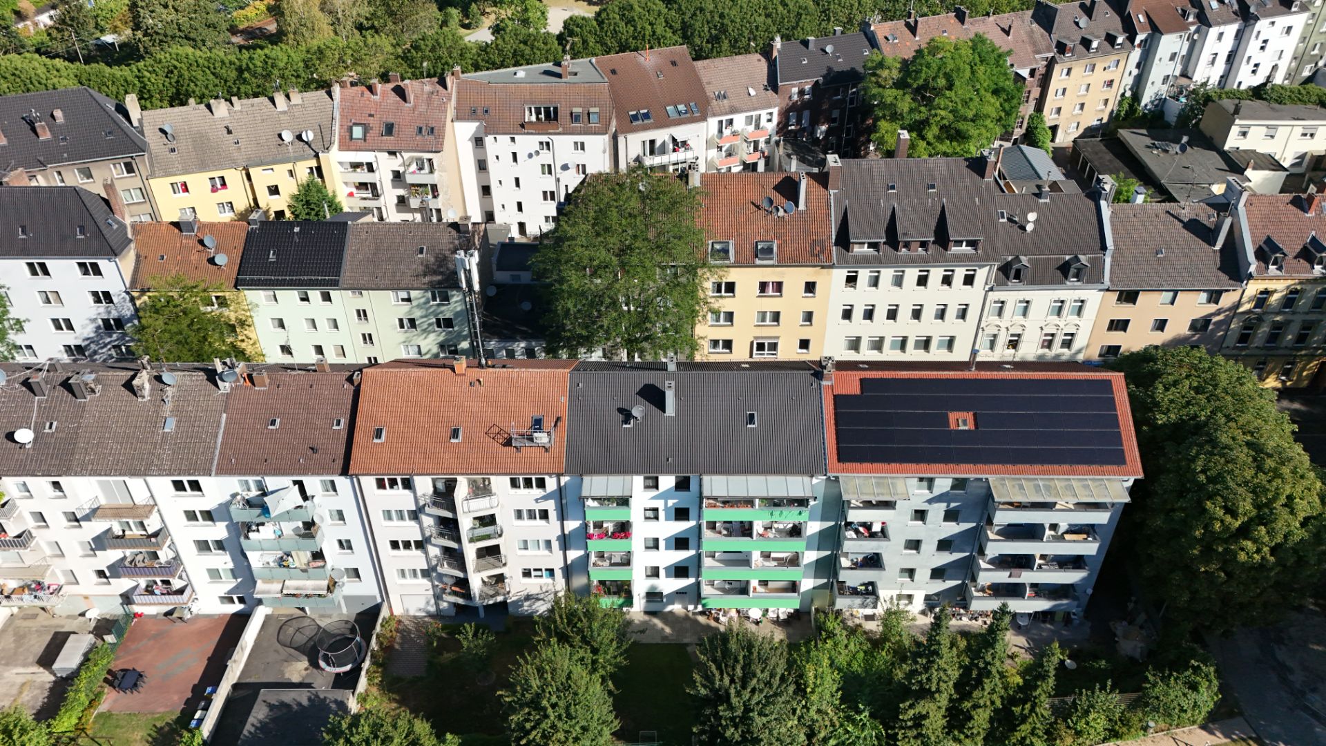 Mehrstöckige Wohnhäuser mit Balkonen und Solarpanels auf einem Dach, Bäume im Vordergrund.