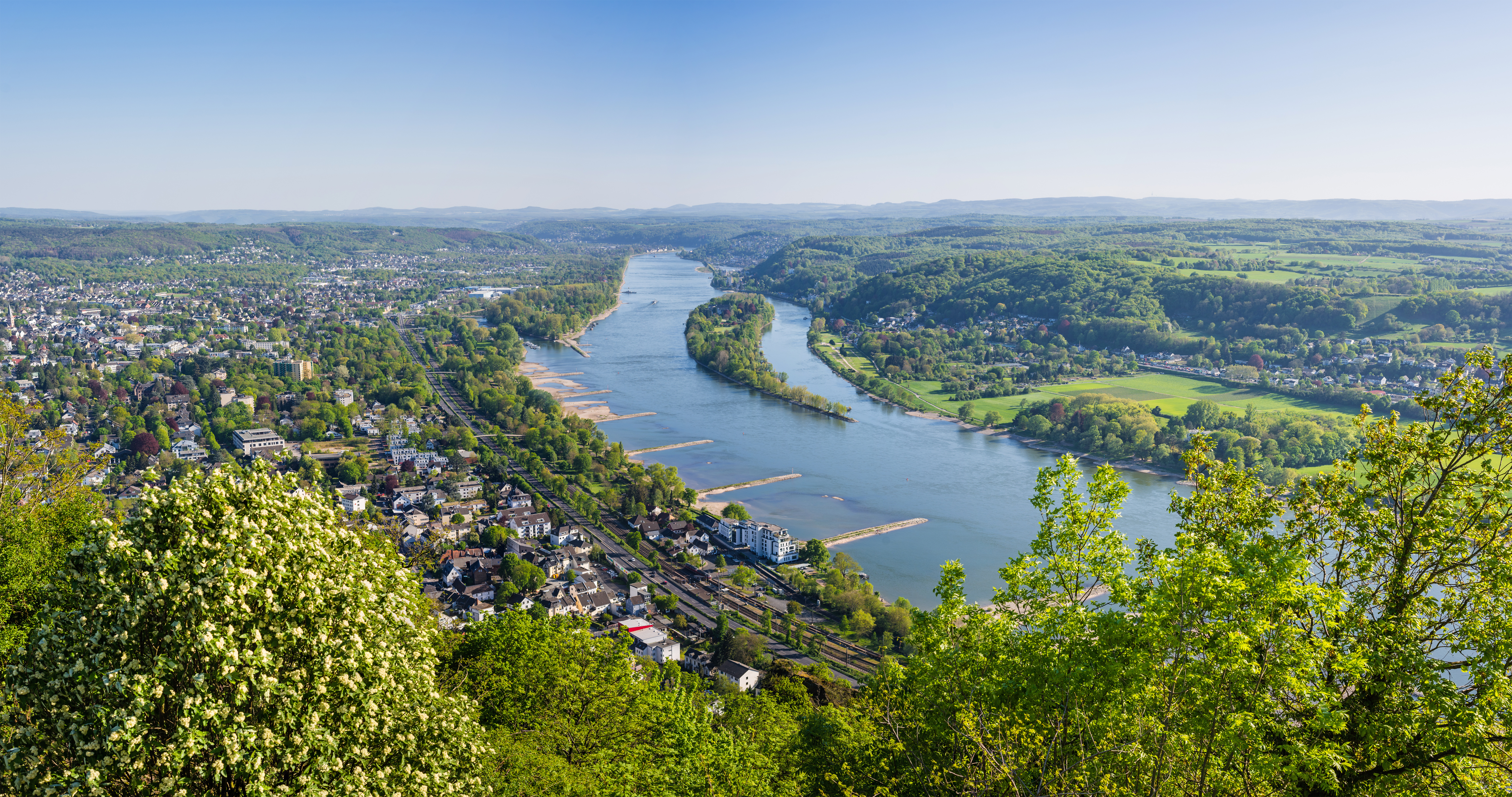 Blick von oben auf einen breiten Fluss, umgeben von grünen Hügeln und Wohnhäusern.