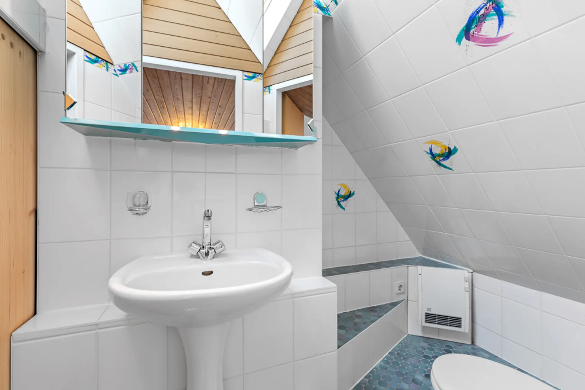 Helles Bad mit Waschbecken, Spiegel, schräger Decke und dekorativen Fliesen an der Wand.
