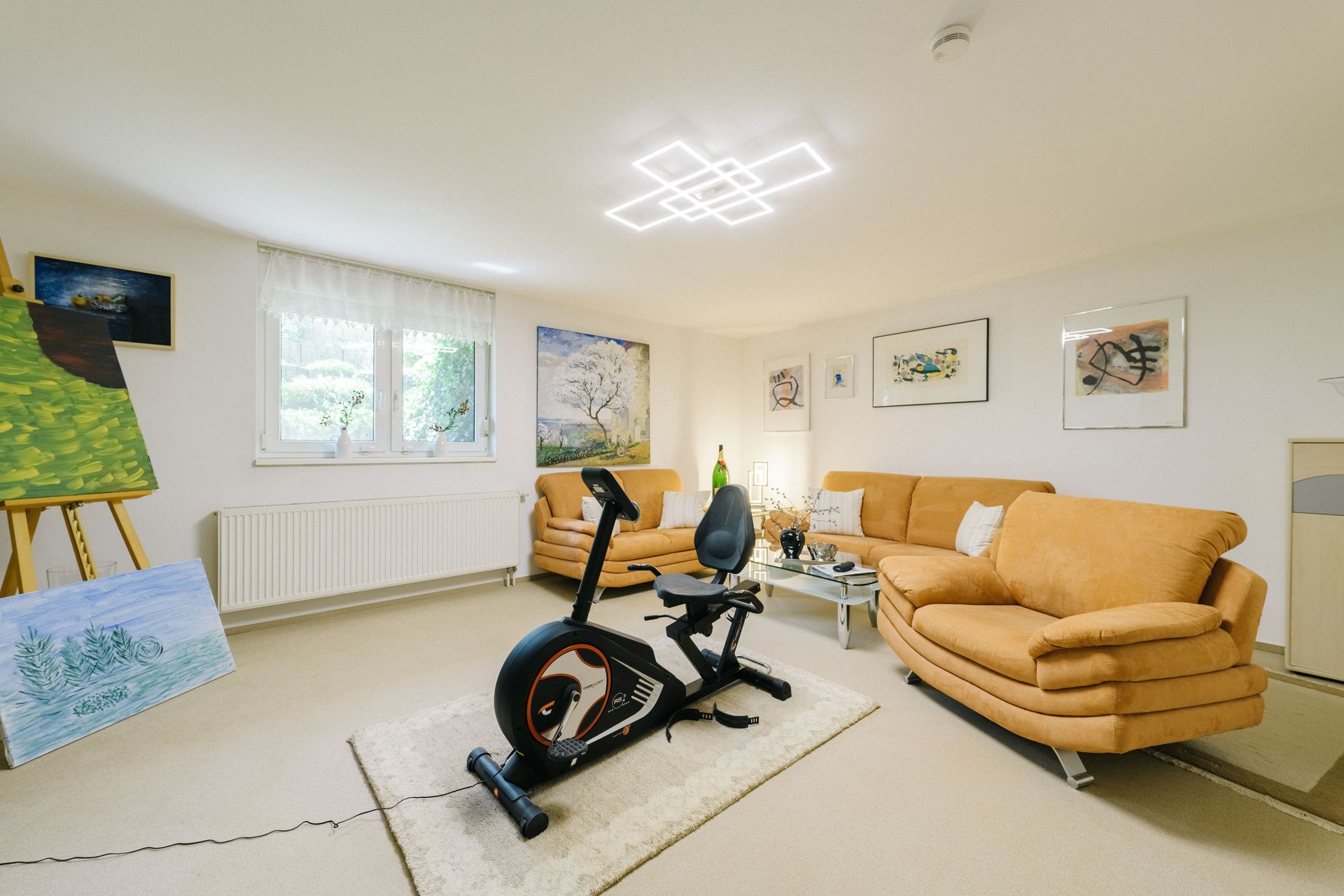 Helles Wohnzimmer mit zwei Sofas, Heimtrainer, mehreren Bildern an den Wänden und Staffeleien mit Gemälden.