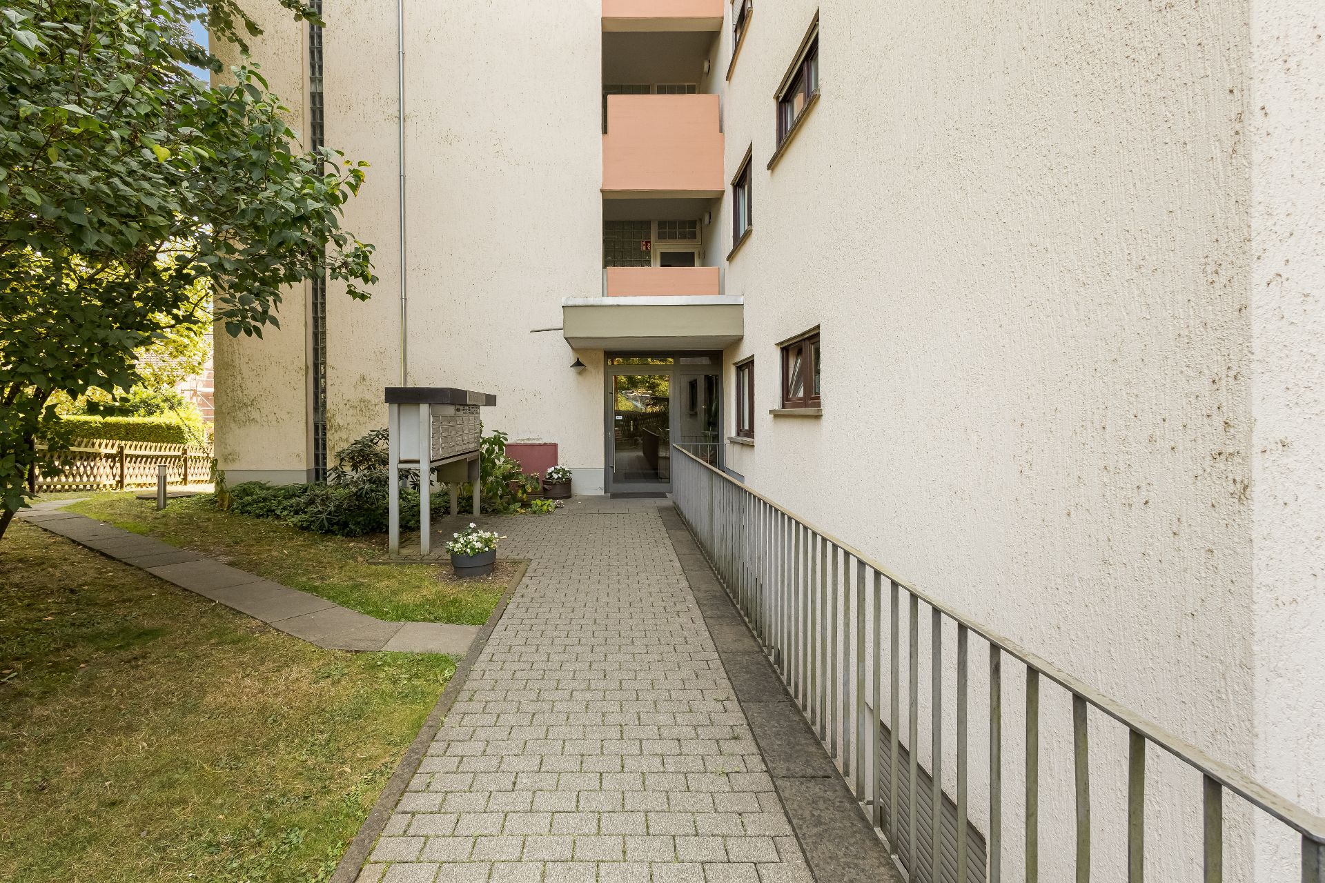 Gepflasterter Weg mit Geländer führt zum Eingang eines mehrstöckigen Wohngebäudes.