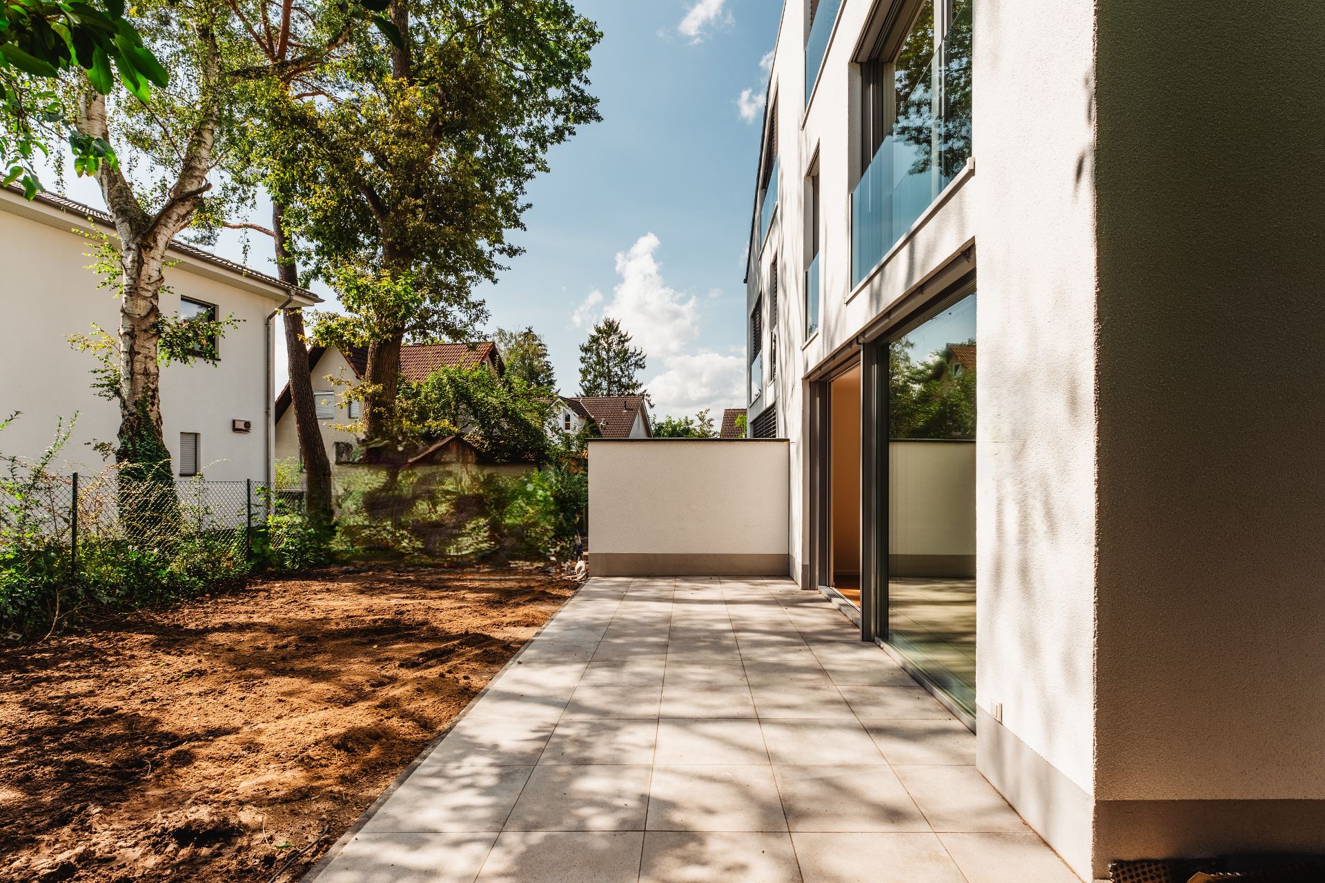 Moderne Terrasse mit Fliesen am Wohnhaus, daneben ein Garten mit Erde und Bäumen.