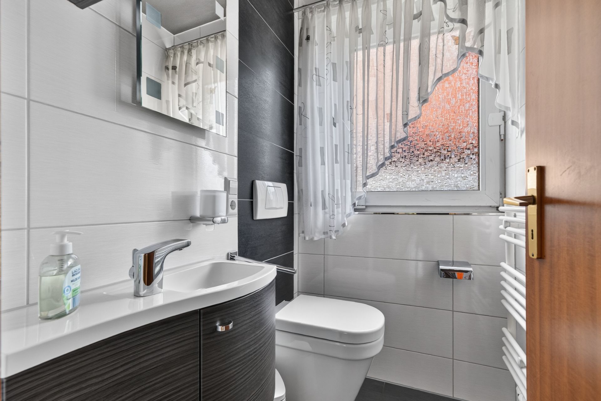 Modernes, kleines Badezimmer mit Waschbecken, WC, Spiegel und Fenster mit Vorhang.