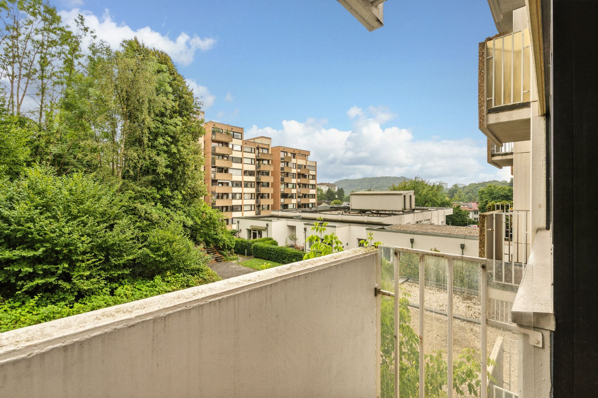 Blick von einem Balkon auf grüne Bäume und ein mehrstöckiges Wohngebäude.