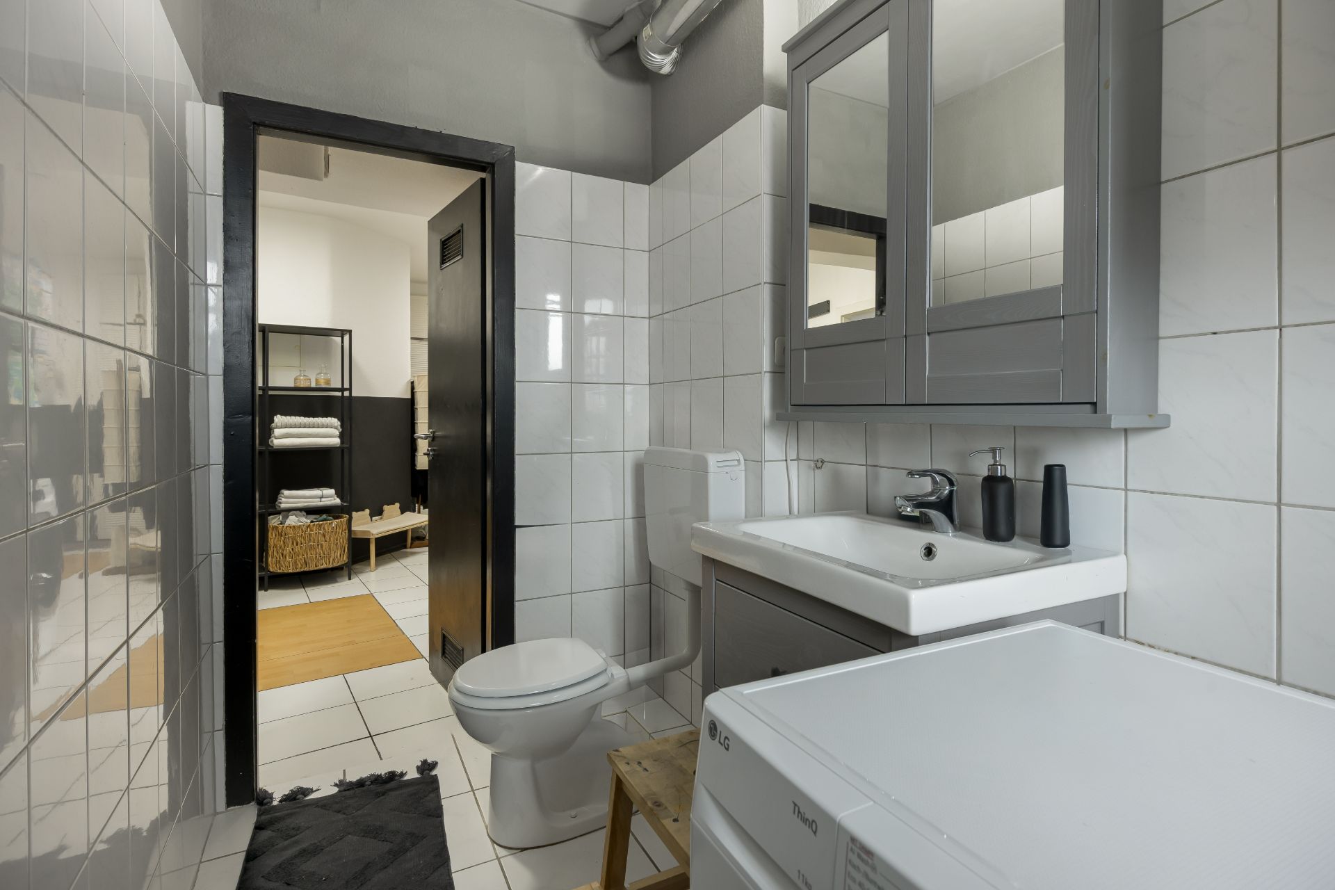 Modernes Badezimmer mit Waschbecken, Toilette, Waschmaschine und Spiegelschrank, Tür zum Flur offen.