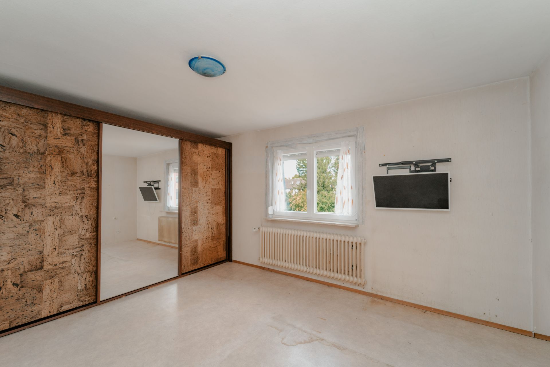 Leeres Zimmer mit großem Fenster, Heizkörper, eingebautem Schrank mit Spiegeltür und Wandhalterung.