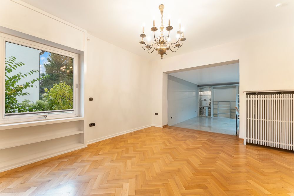 Helles Wohnzimmer mit Parkettboden, großem Fenster, Kronleuchter und offener Verbindung zum nächsten Raum.