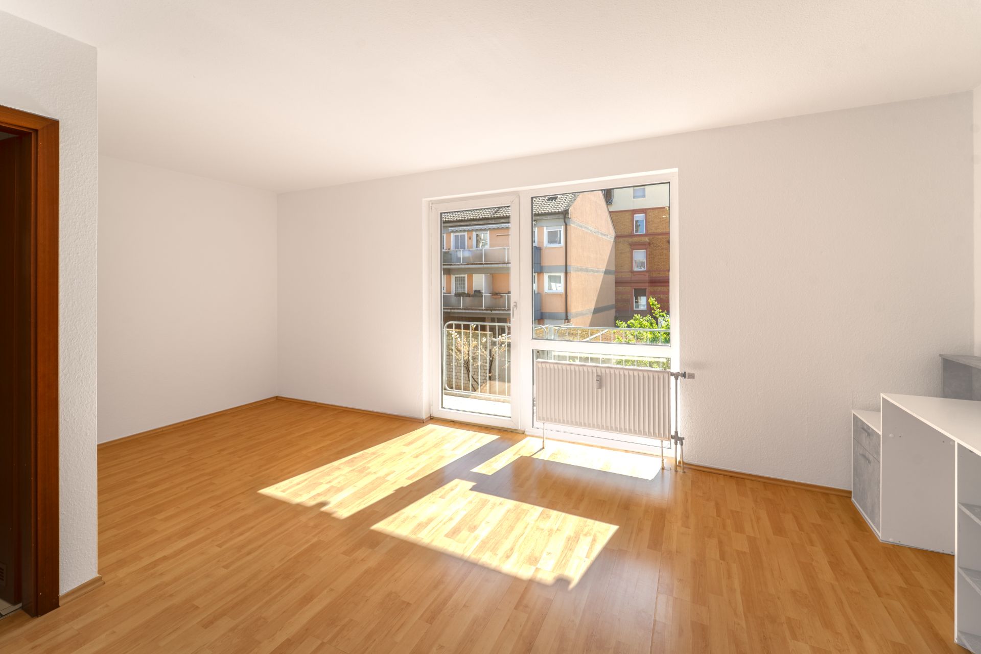 Helles, leeres Zimmer mit Parkettboden und großem Fenster zur Straße.