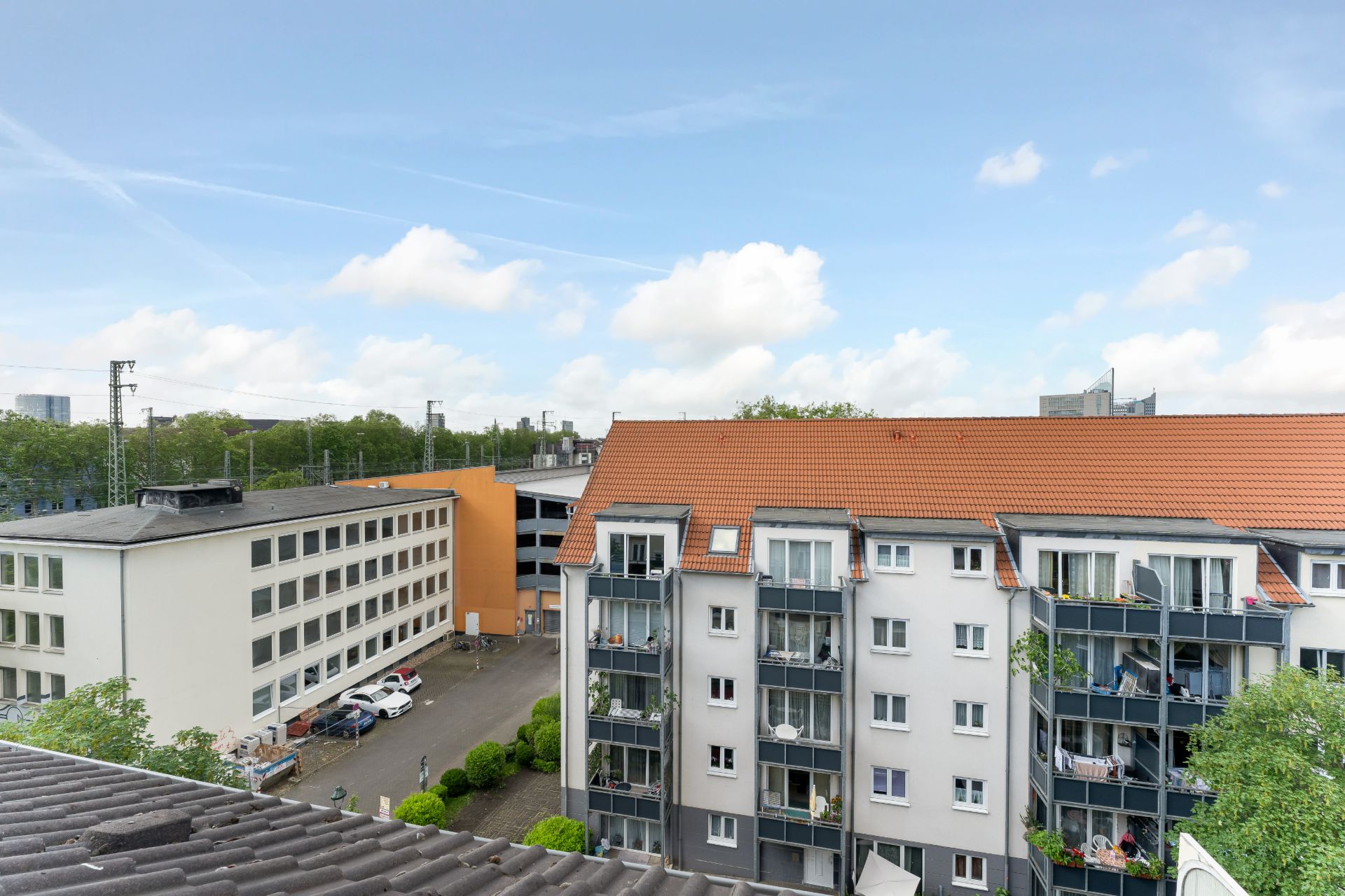 Mehrstöckiges Wohngebäude mit Balkonen und rotem Dach, daneben ein Bürogebäude und geparkte Autos.