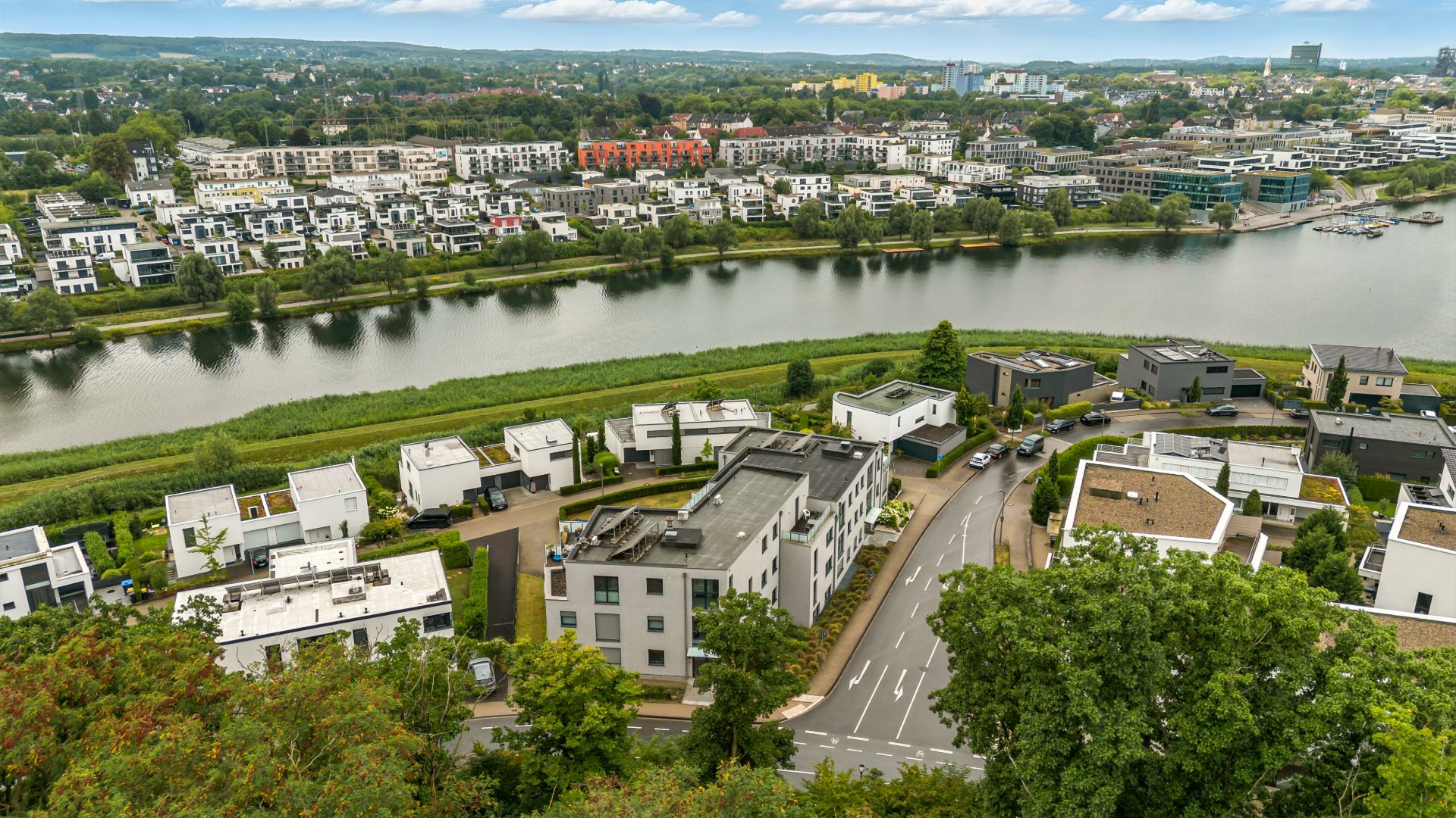 Luftaufnahme von Wohnhäusern an einem Fluss mit Straßen, Bäumen und weiteren Gebäuden im Hintergrund.