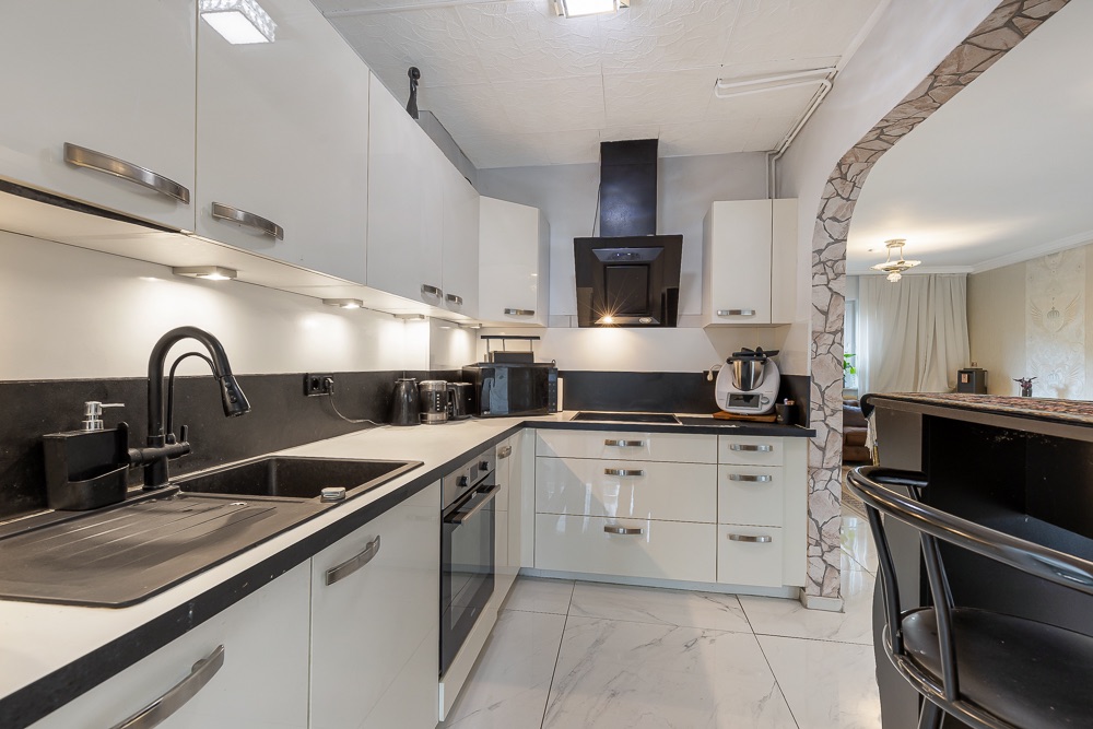 Moderne Einbauküche mit weißen Schränken, Geräten und Bartresen, angrenzend an ein Wohnzimmer.
