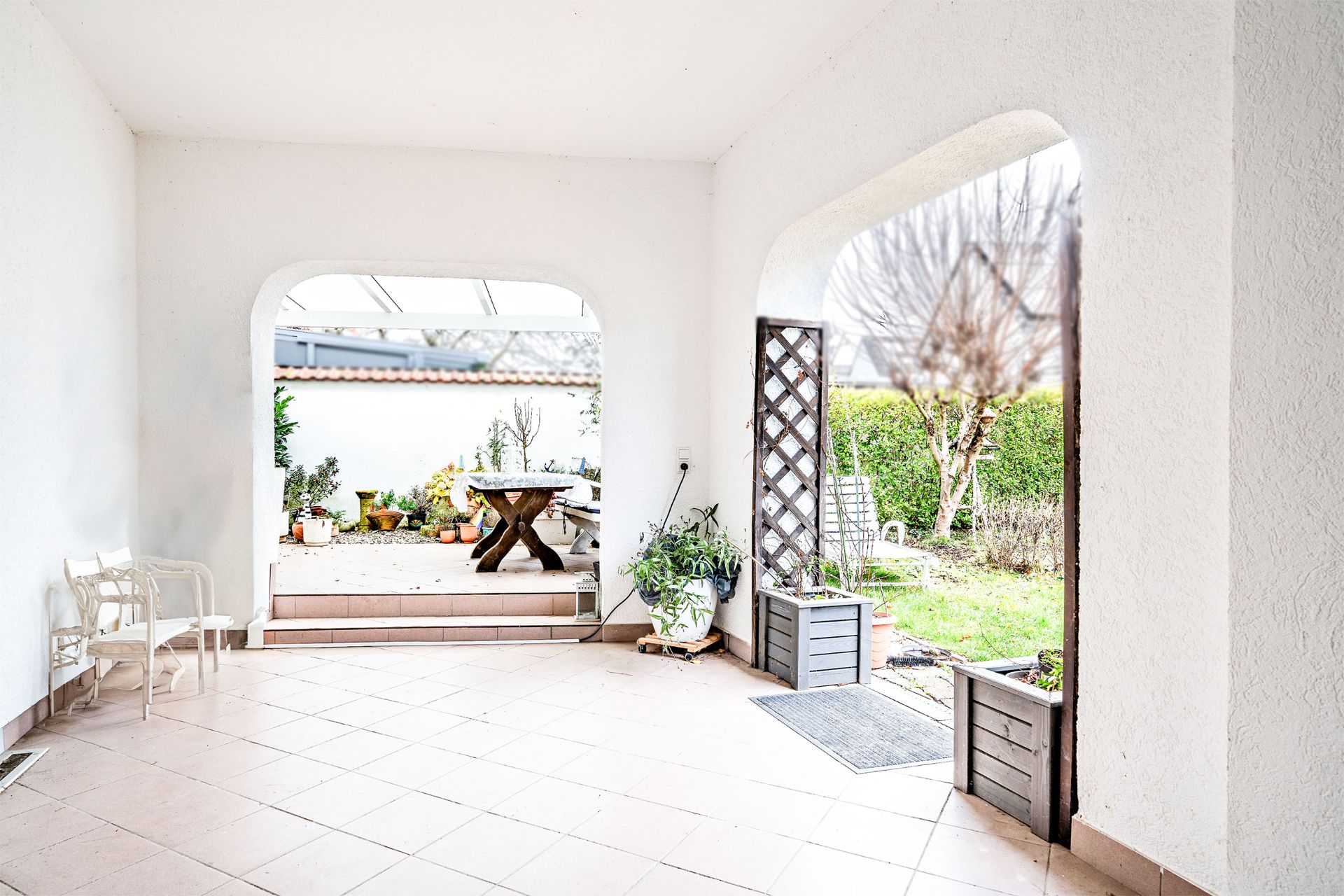 Überdachte Terrasse mit Fliesenboden, einigen Stühlen und Blick in einen Garten mit Pflanzen.