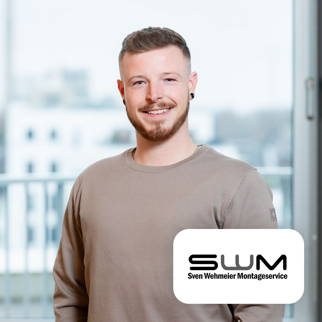 Ein junger Mann steht lächelnd in einem hellen Raum mit Balkon im Hintergrund.