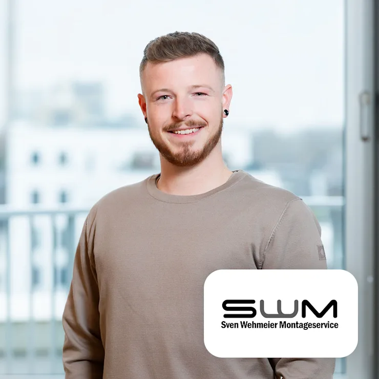 Ein junger Mann steht lächelnd in einem hellen Raum mit Balkon im Hintergrund.