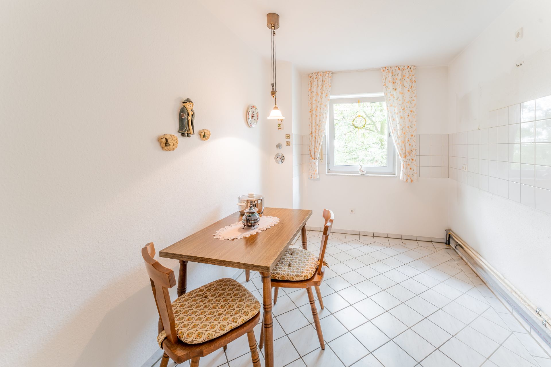 Heller Raum mit Esstisch, zwei Stühlen, Fenster mit Vorhängen und Wanddekorationen.