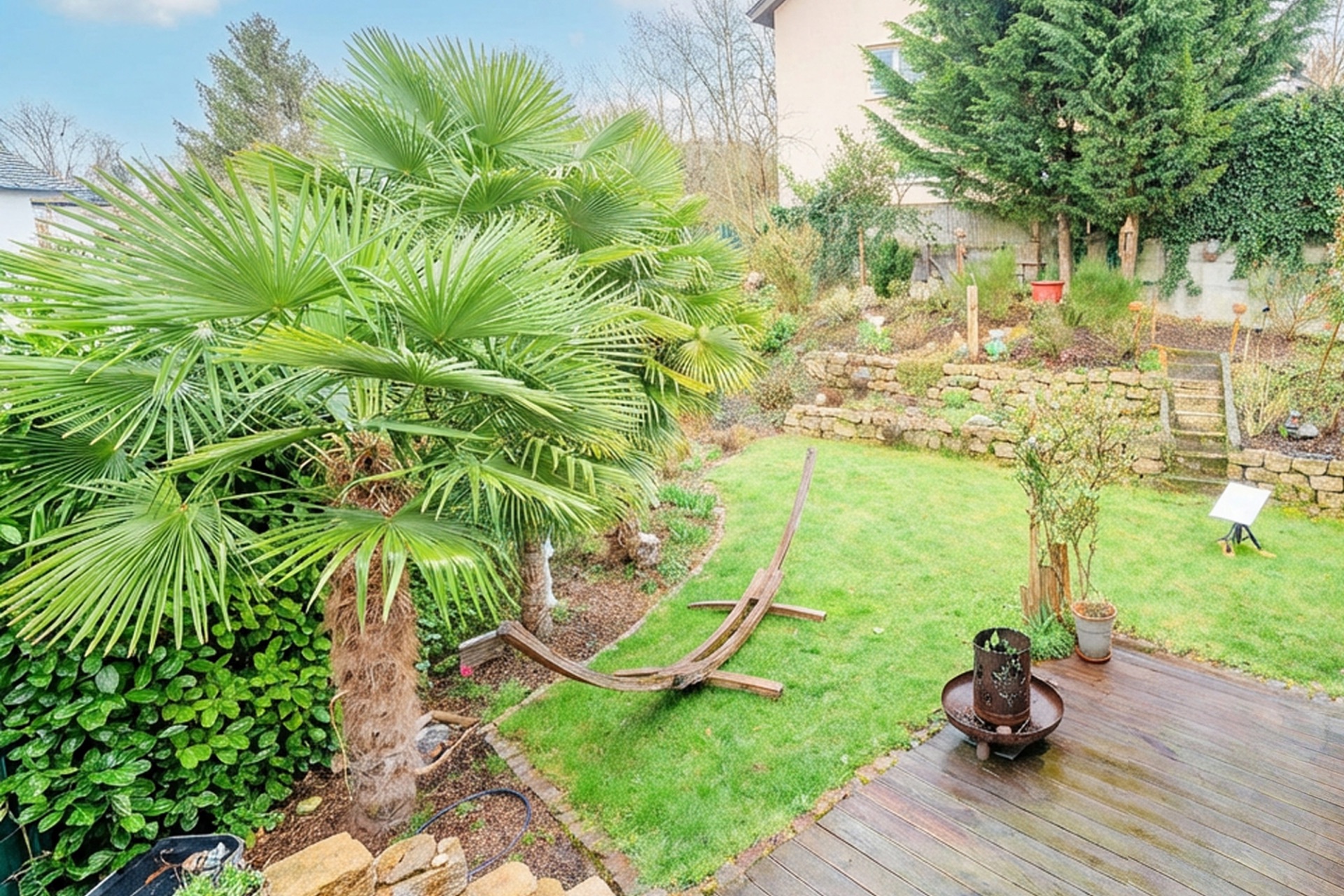 Garten mit Palmen, Holzterrasse, Feuerstelle und leerem Hängemattengestell.