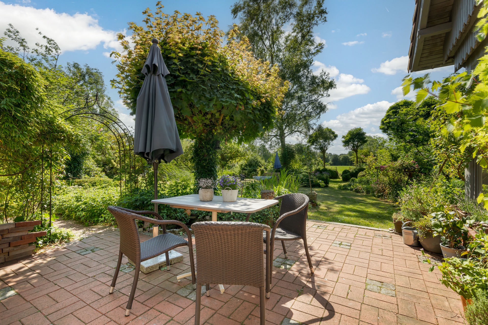 Gepflasterte Terrasse mit Tisch, vier Stühlen und Sonnenschirm, umgeben von Garten und Bäumen.