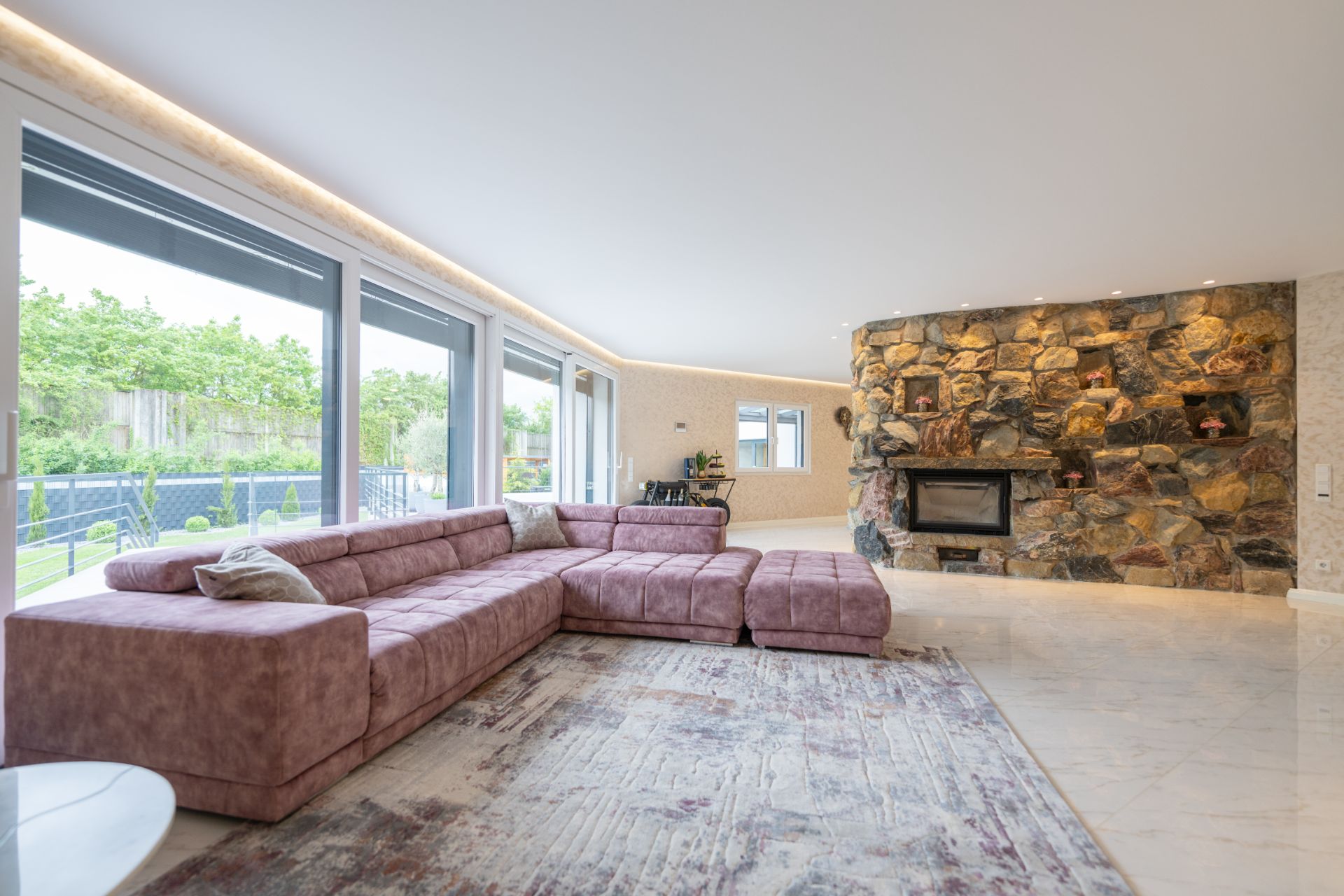 Modernes Wohnzimmer mit großer Eckcouch, Steinwand mit Kamin und großen Fenstern zum Garten.