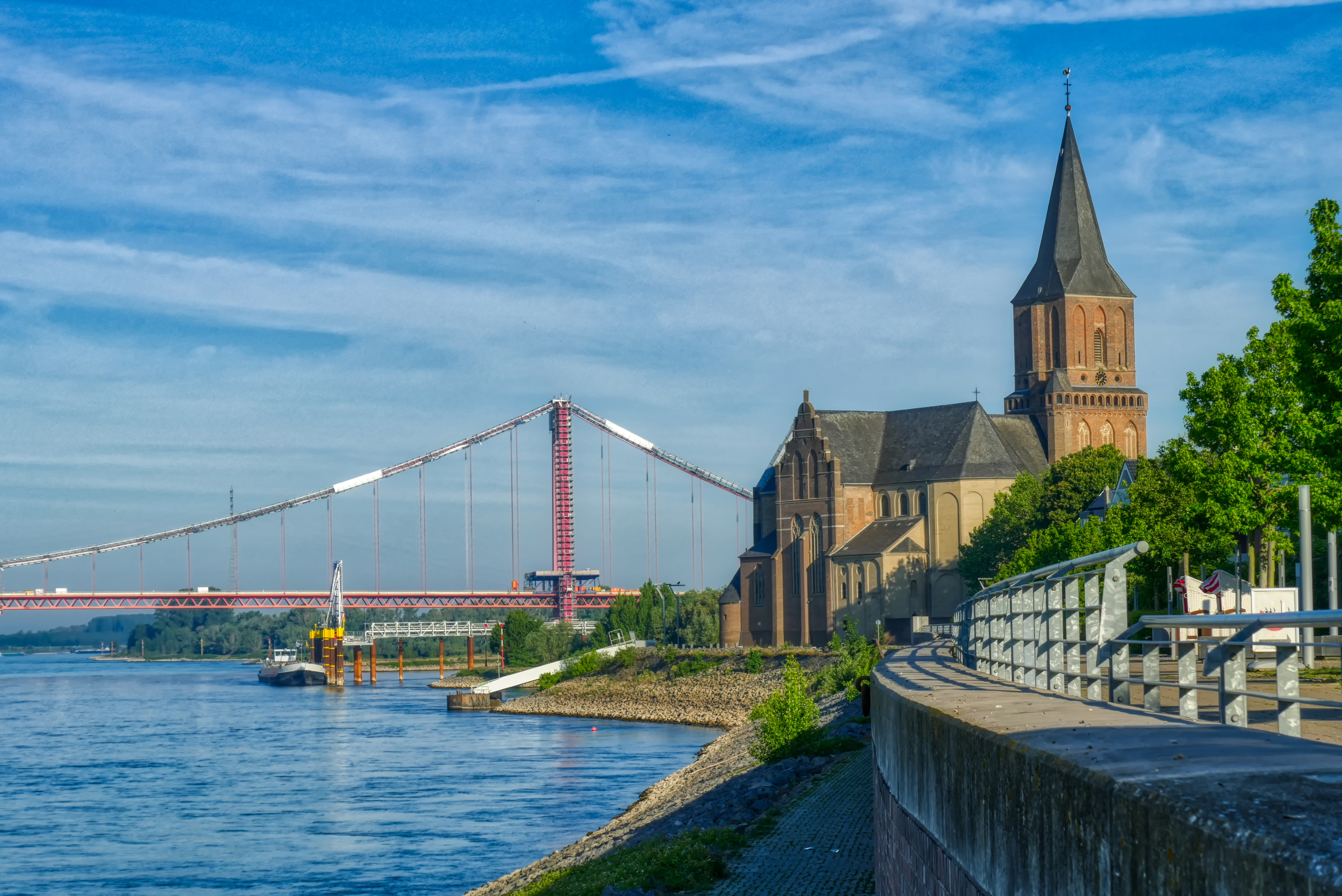 Flussufer mit Kirche, Bäumen, Gehweg und einer Brücke im Hintergrund an einem sonnigen Tag.