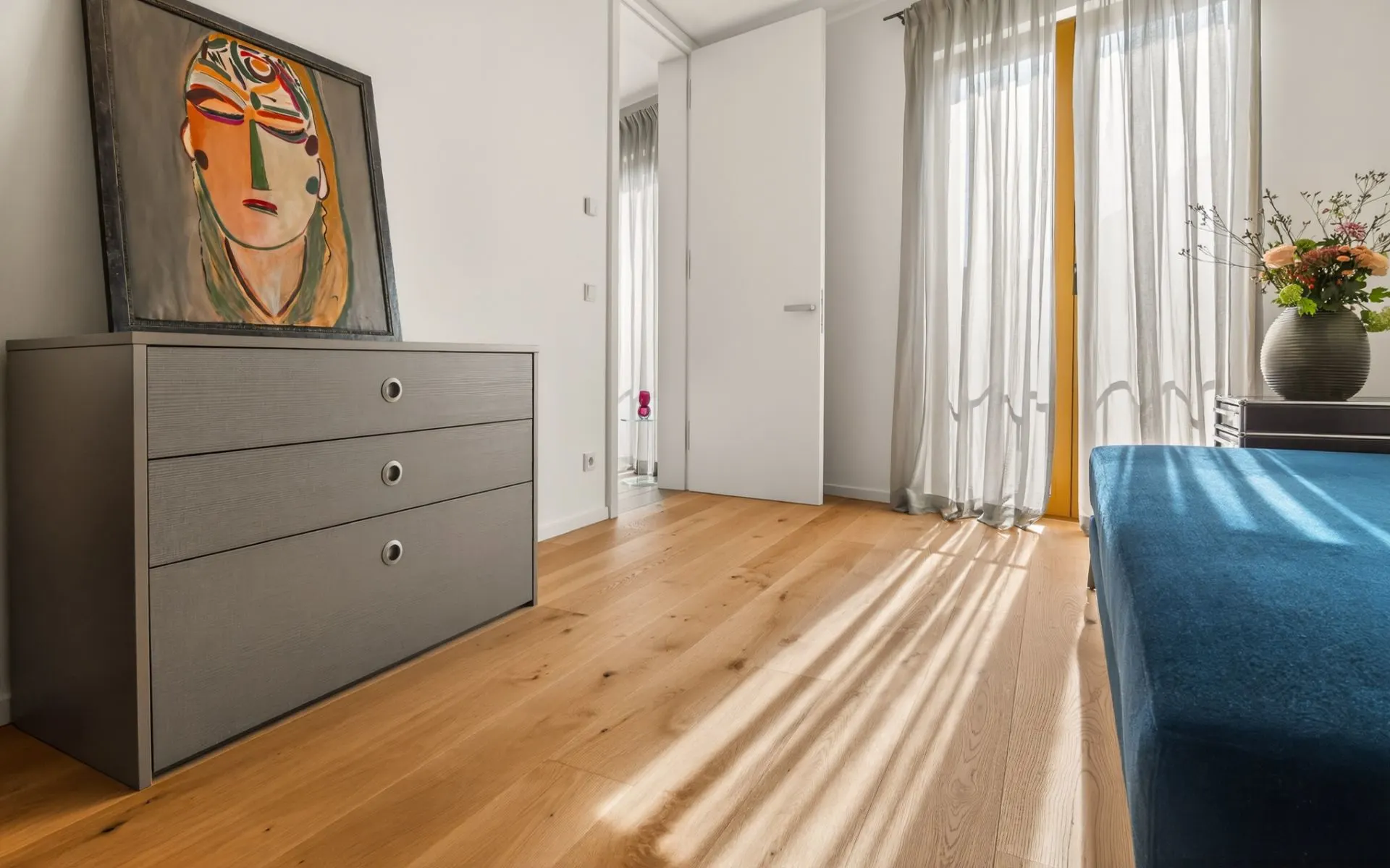 Helles Zimmer mit Kommode, Bild, blauem Sofa und Vorhängen vor Fenstern.