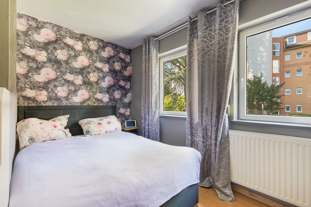 Schlafzimmer mit Doppelbett, floraler Tapete und großen Fenstern mit grauen Vorhängen.