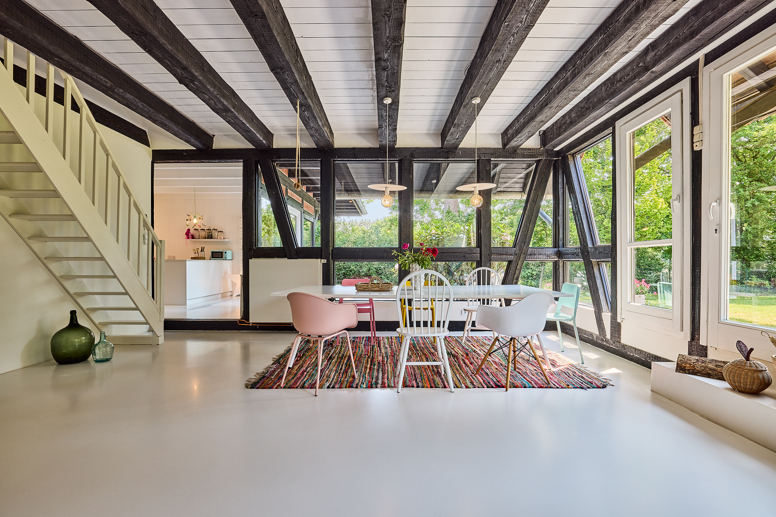 Heller Essbereich mit bunten Stühlen, Holzbalken, großem Fenster und Treppe im Hintergrund.