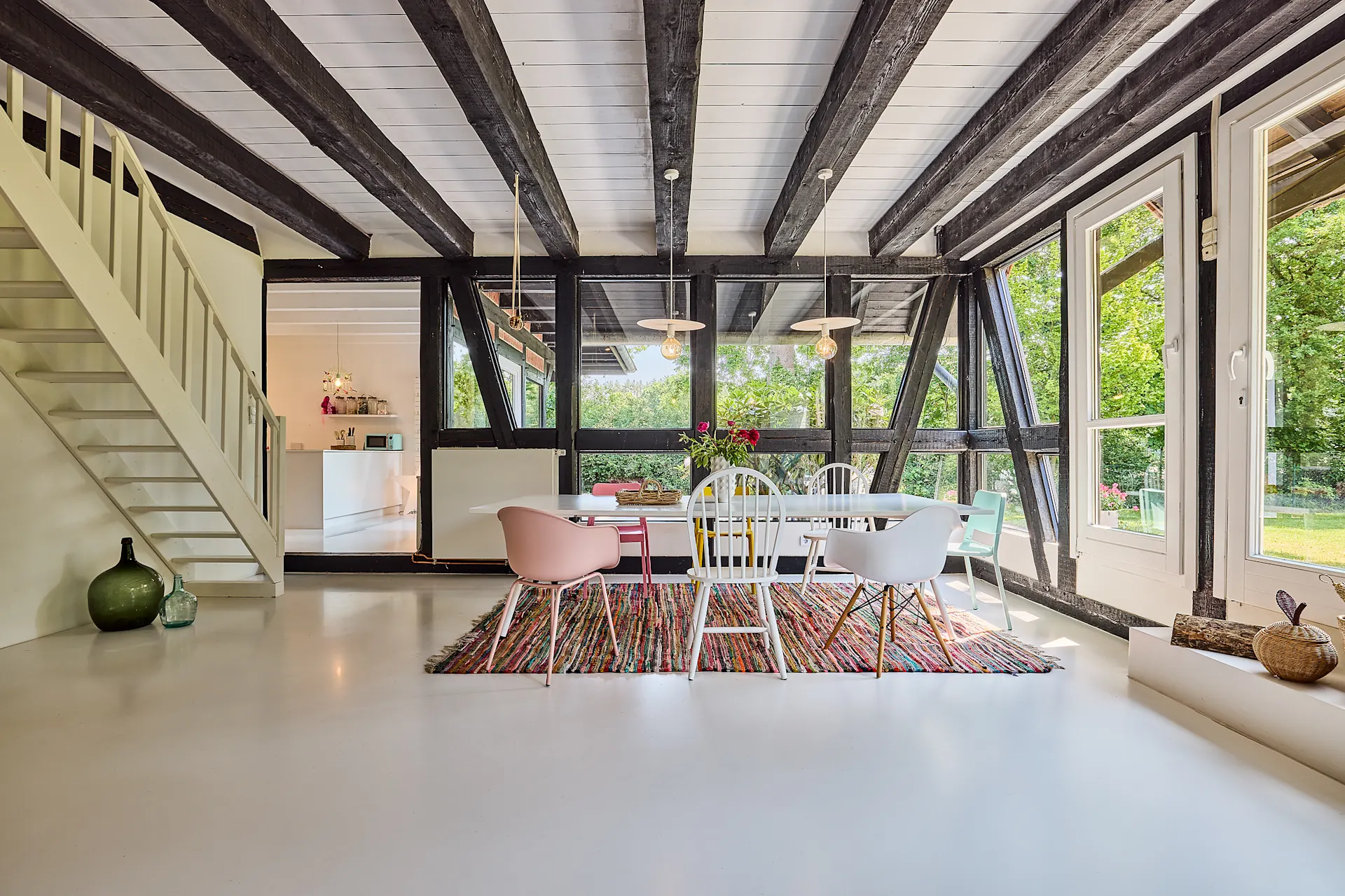 Heller Essbereich mit bunten Stühlen, Holzbalken, großem Fenster und Treppe im Hintergrund.