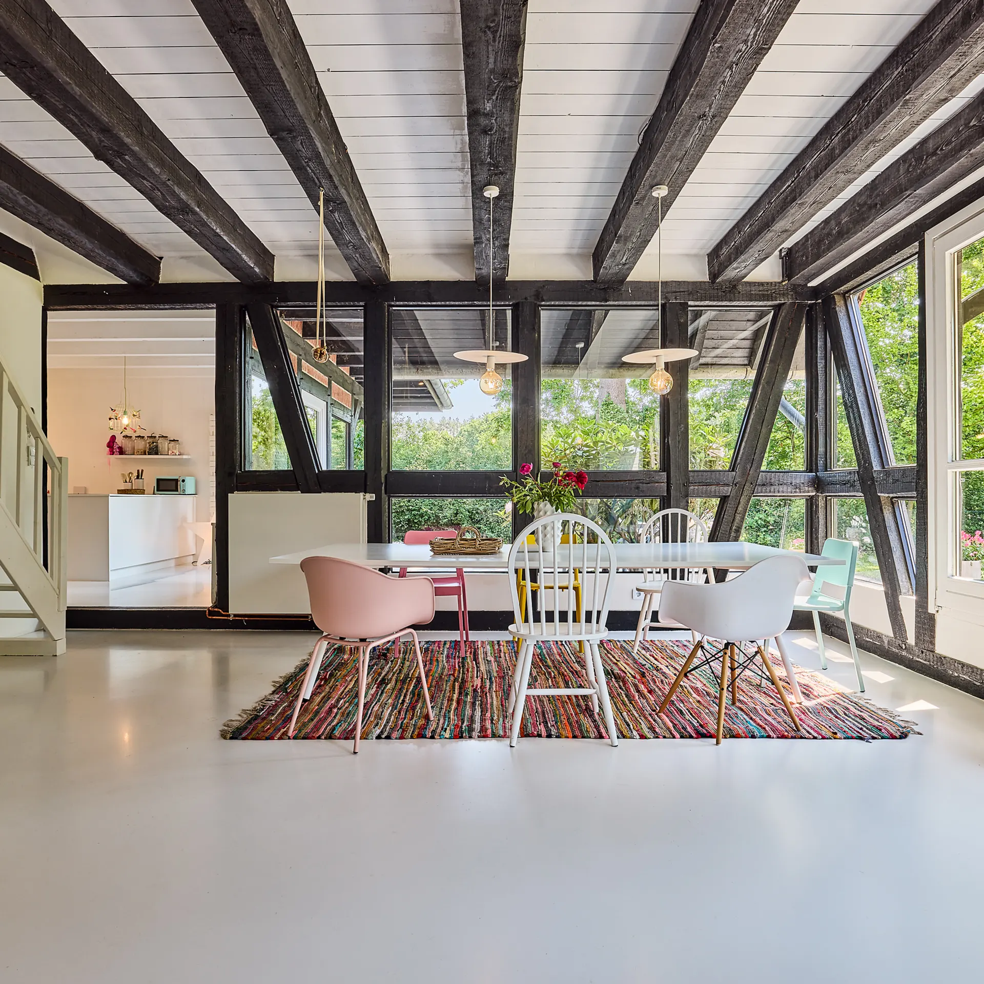 Heller Essbereich mit bunten Stühlen, Holzbalken, großem Fenster und Treppe im Hintergrund.