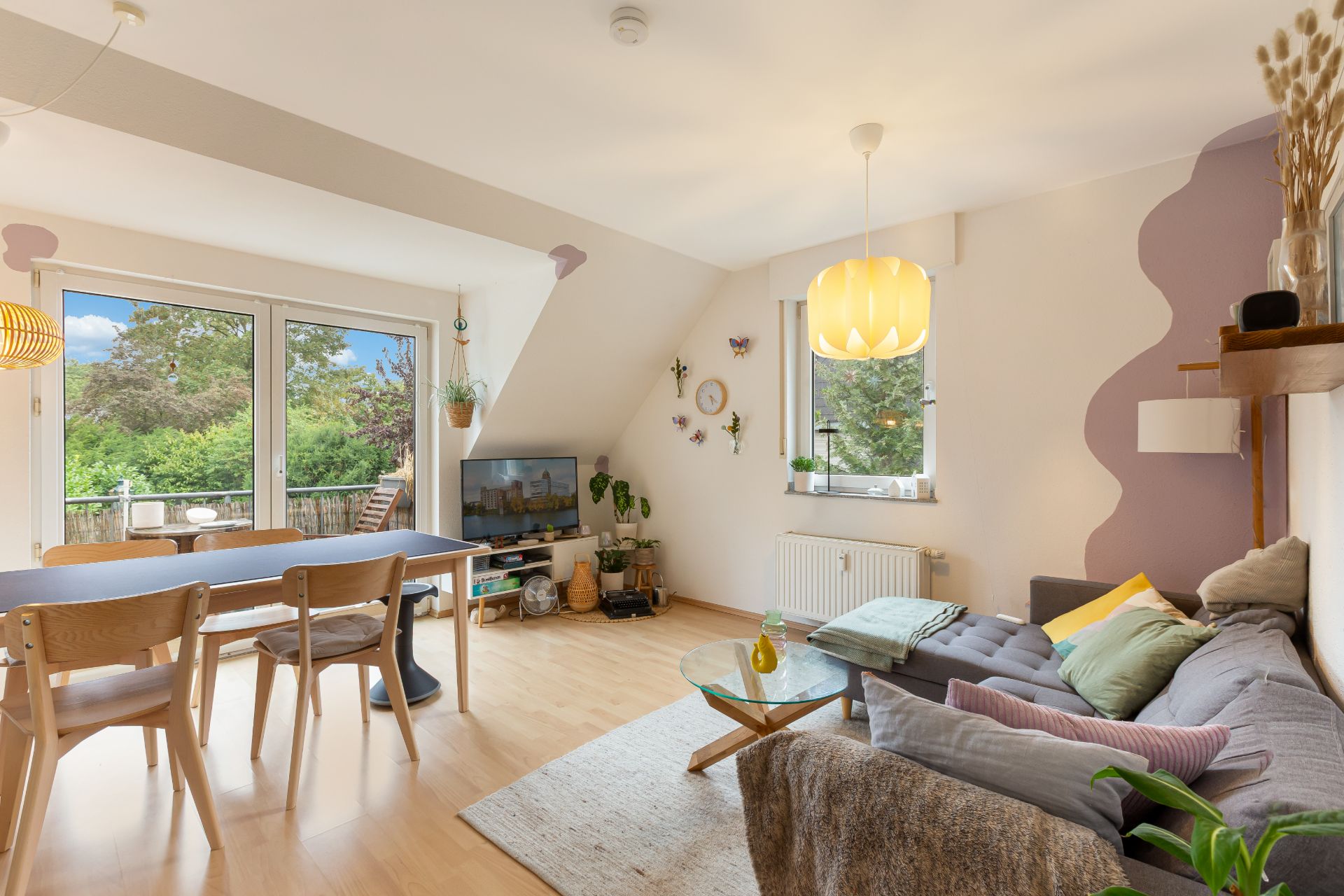 Helles Wohnzimmer mit Sofa, Esstisch, Fernseher und großen Fenstern mit Blick auf den Garten.