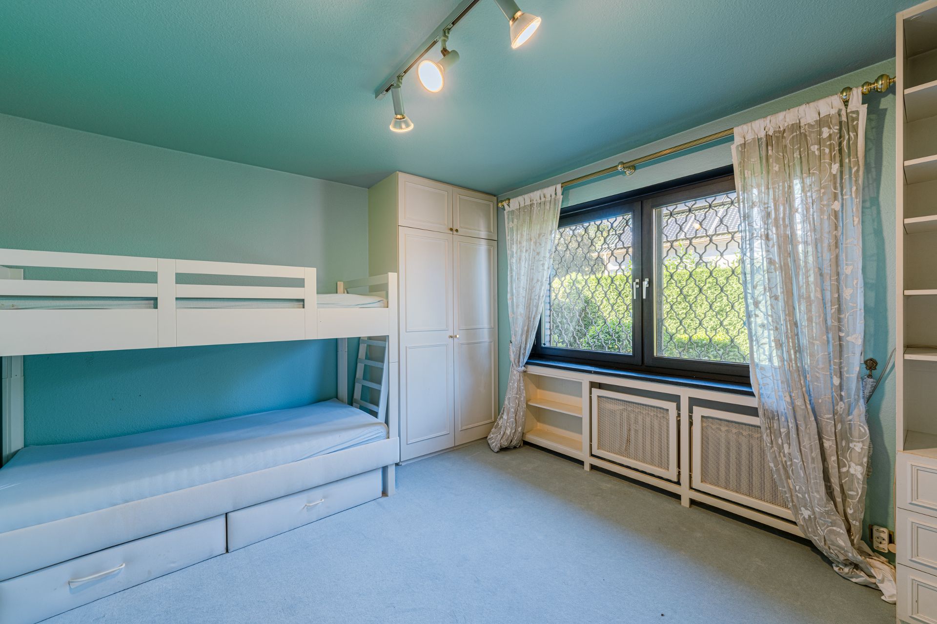 Helles Zimmer mit Etagenbett, großem Fenster, Vorhängen und weißen Schränken.