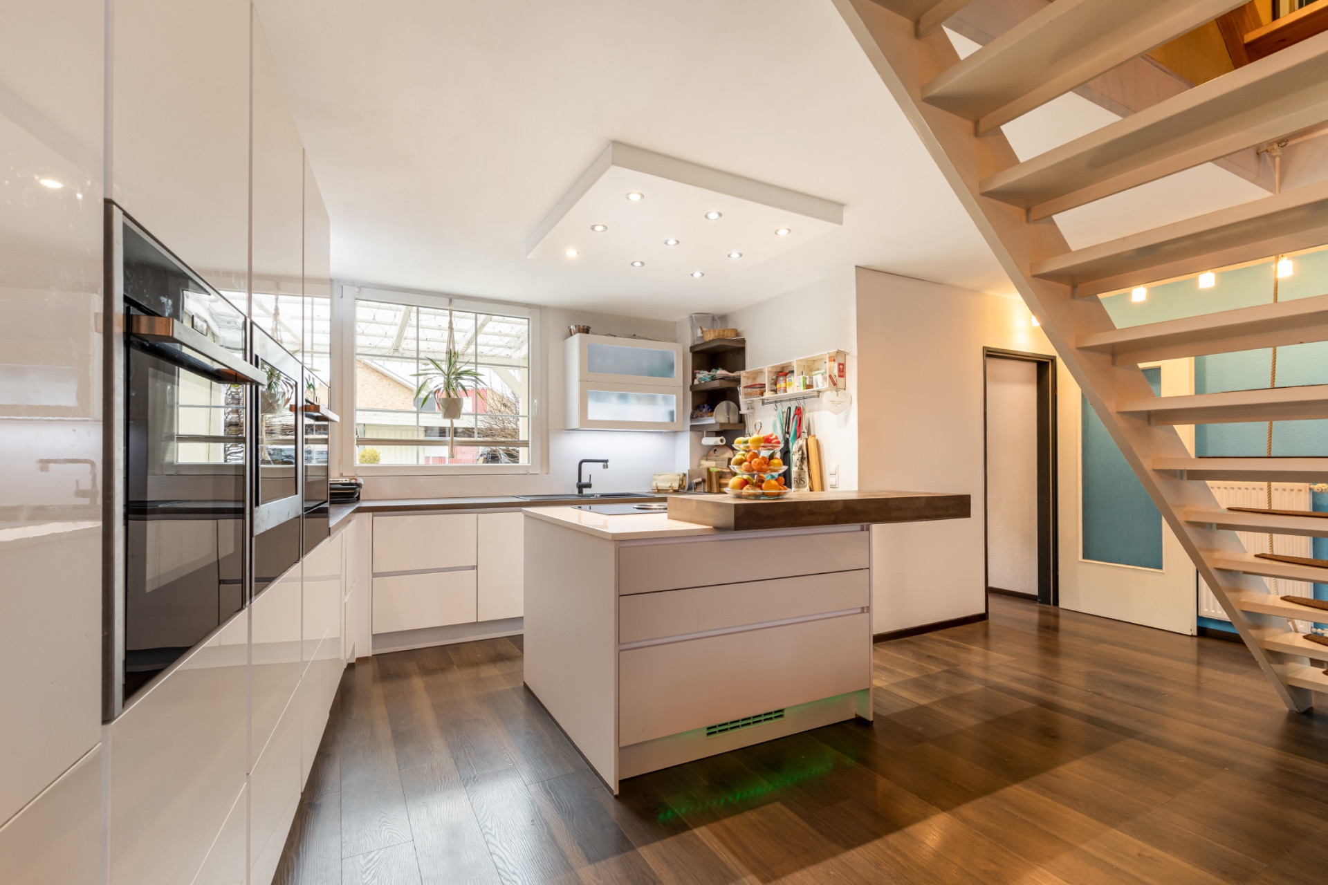 Moderne Küche mit Kochinsel, Holzboden und Treppe, großes Fenster im Hintergrund.