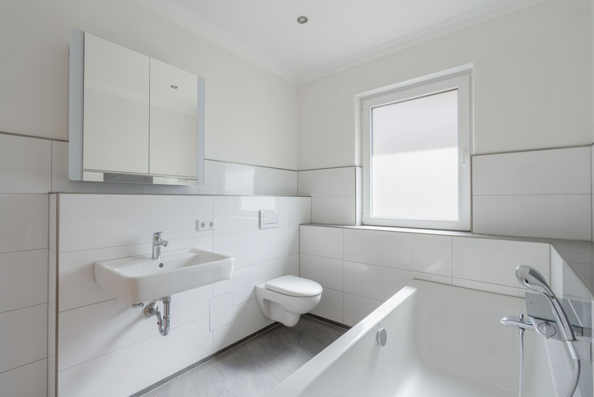 Helles Badezimmer mit Badewanne, Wandwaschbecken, Spiegel und Fenster.