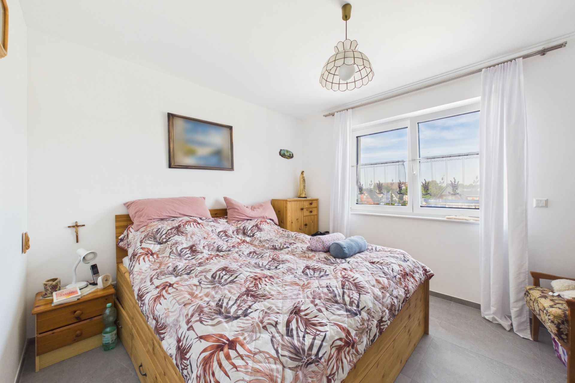 Helles Schlafzimmer mit Doppelbett, zwei Nachttischen, Holzkommode und Fenster mit weißen Vorhängen.