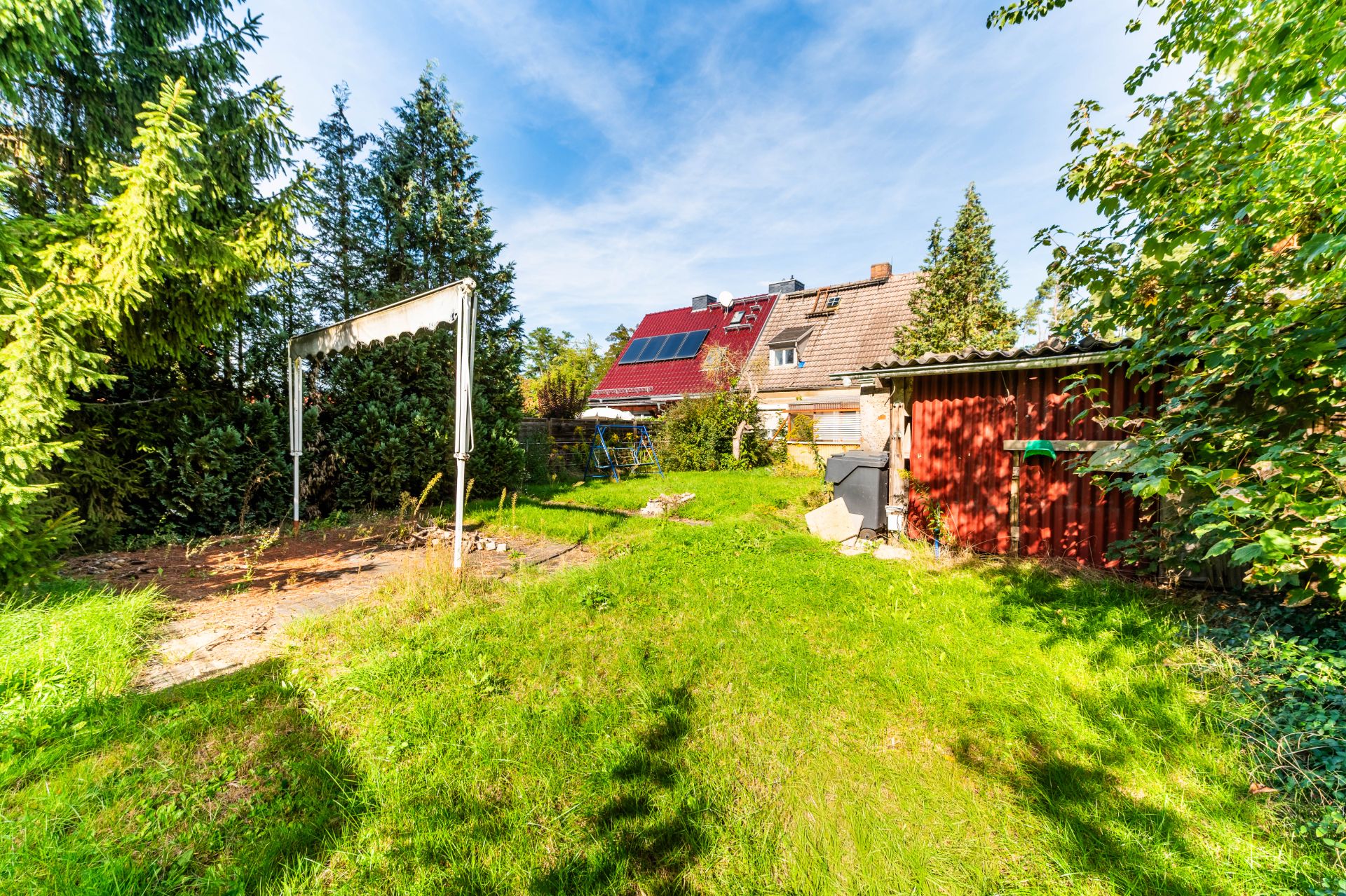 Grüner Garten mit Rasen, Bäumen, Schuppen und einem Haus mit Solarpanels im Hintergrund.