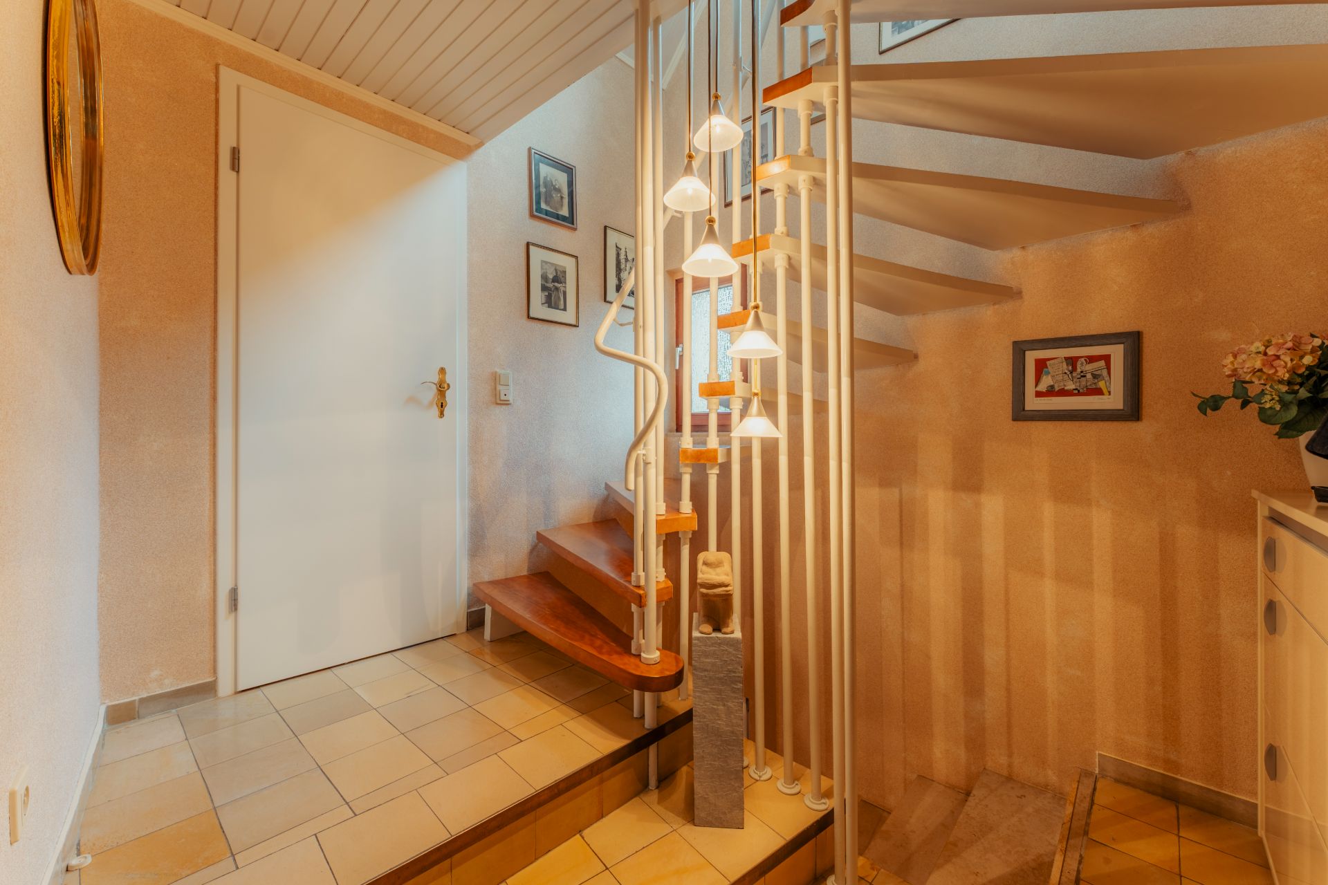 Treppenhaus mit Holztreppe, weißen Geländern, heller Tür und dekorativen Bildern an der Wand.