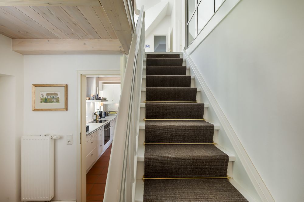 Treppe mit dunklem Teppich, daneben Zugang zu einer Küche, helle Wände und Holzdecke.