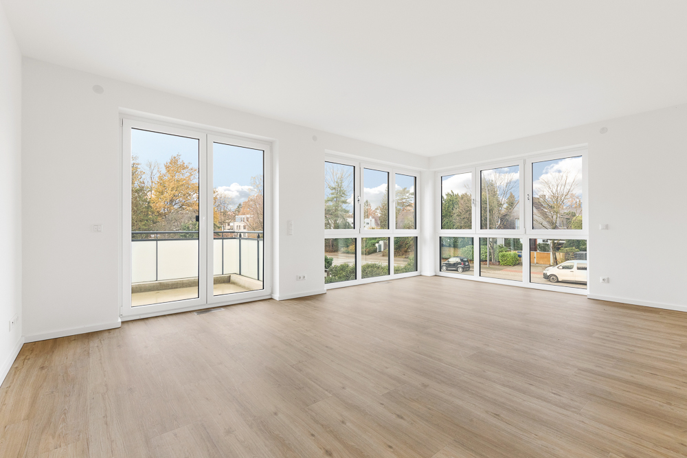Helles, leeres Wohnzimmer mit großen Fenstern und Zugang zum Balkon, Parkettboden.