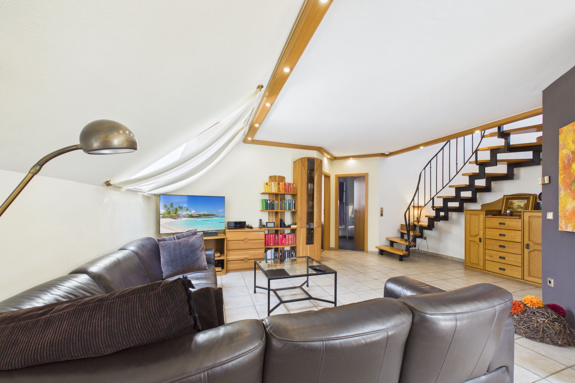 Helles Wohnzimmer mit Ledersofa, Fernseher, Bücherregal und offener Treppe.