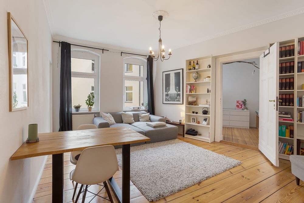 Helles Wohnzimmer mit Sofa, Teppich, Esstisch, Regalen und Zugang zu weiterem Raum.